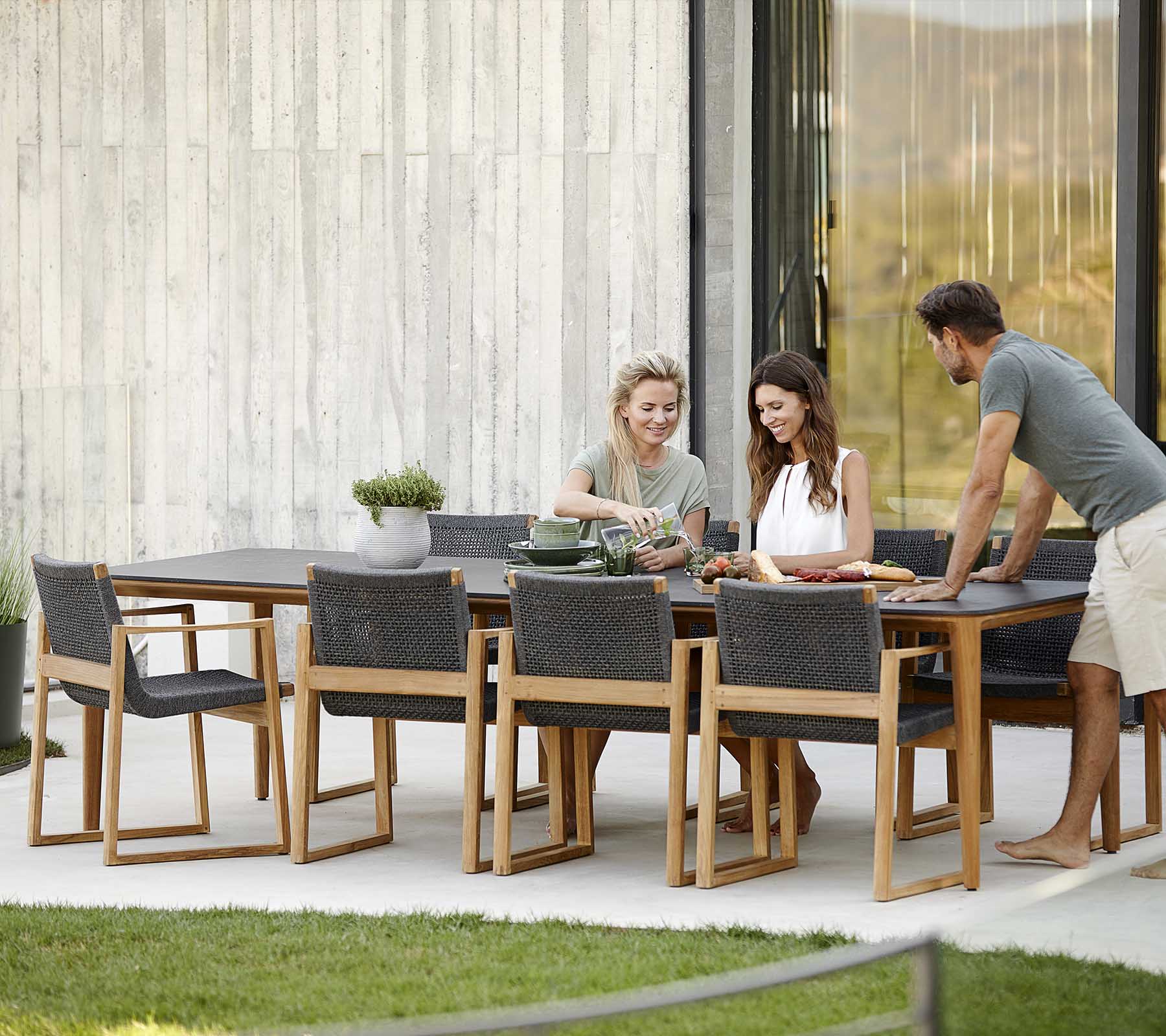 Cuatro personas disfrutan de una comida juntas en un elegante juego de comedor de madera, con sillas modernas y un entorno al aire libre sereno.
