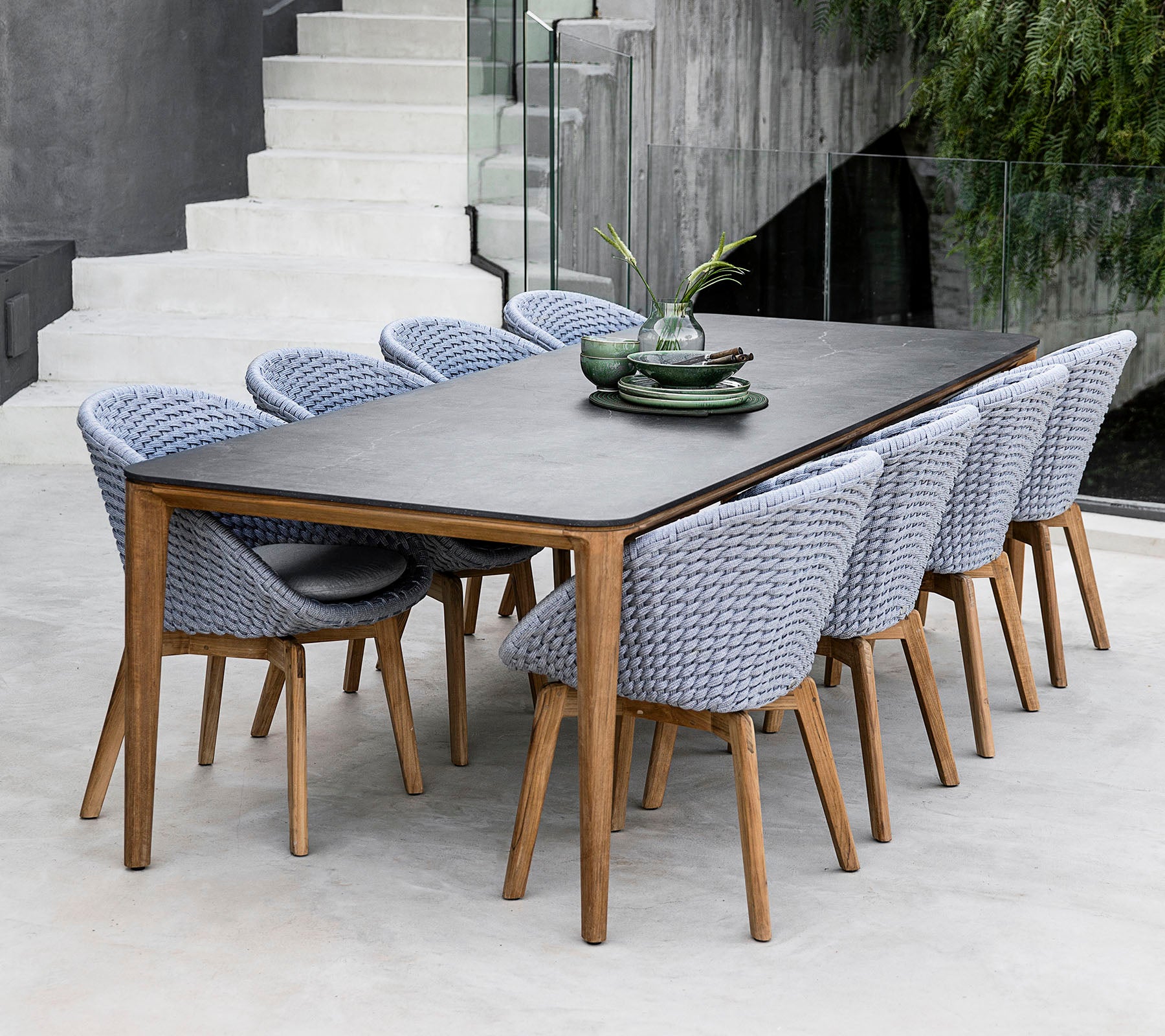Una mesa de comedor elegante con una superficie oscura, acompañada de sillas trenzadas de color claro, complementada con platos decorativos y follaje.