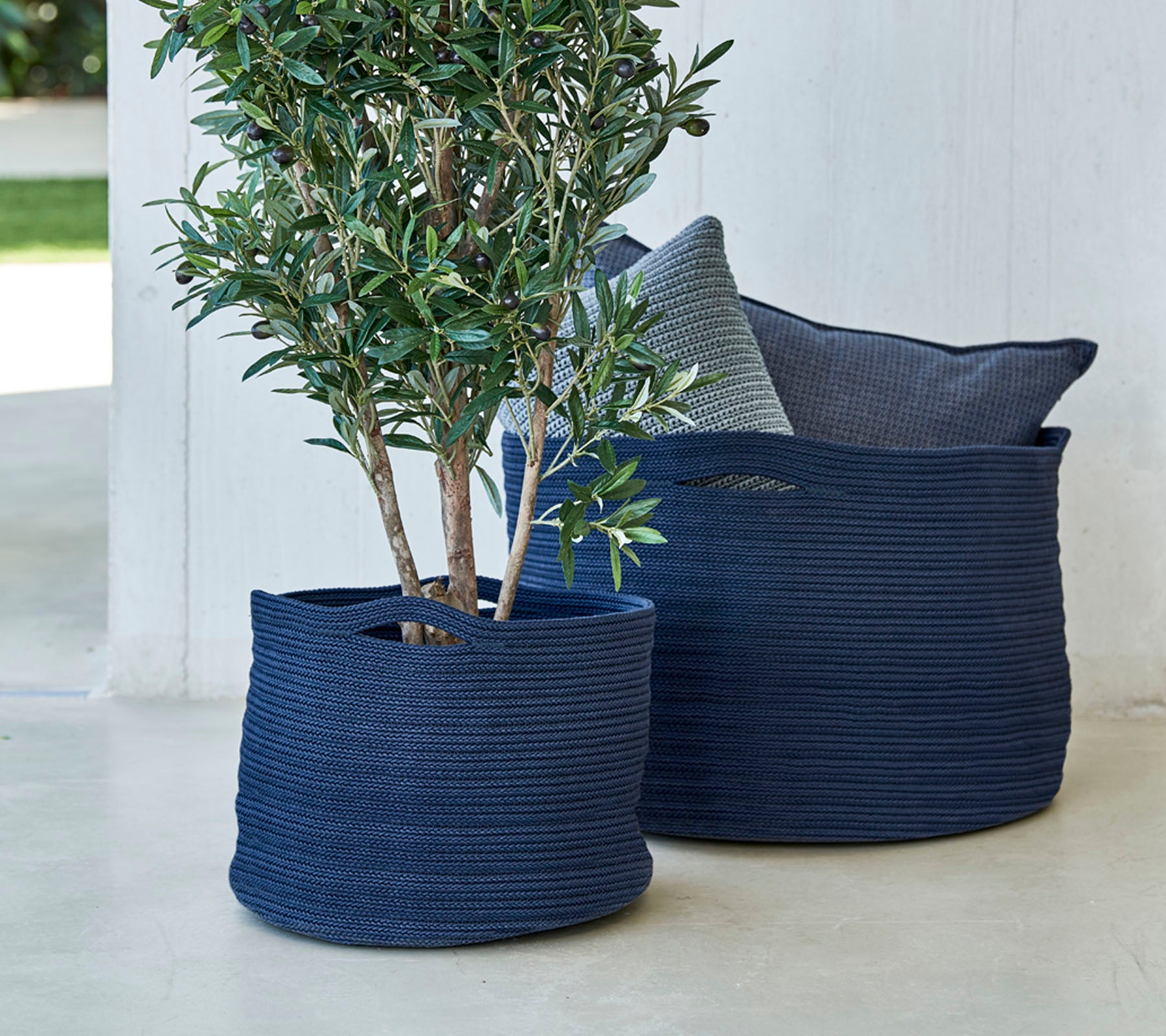Dos macetas tejidas de color azul marino, una con un pequeño árbol, acompañadas de un cojín decorativo.