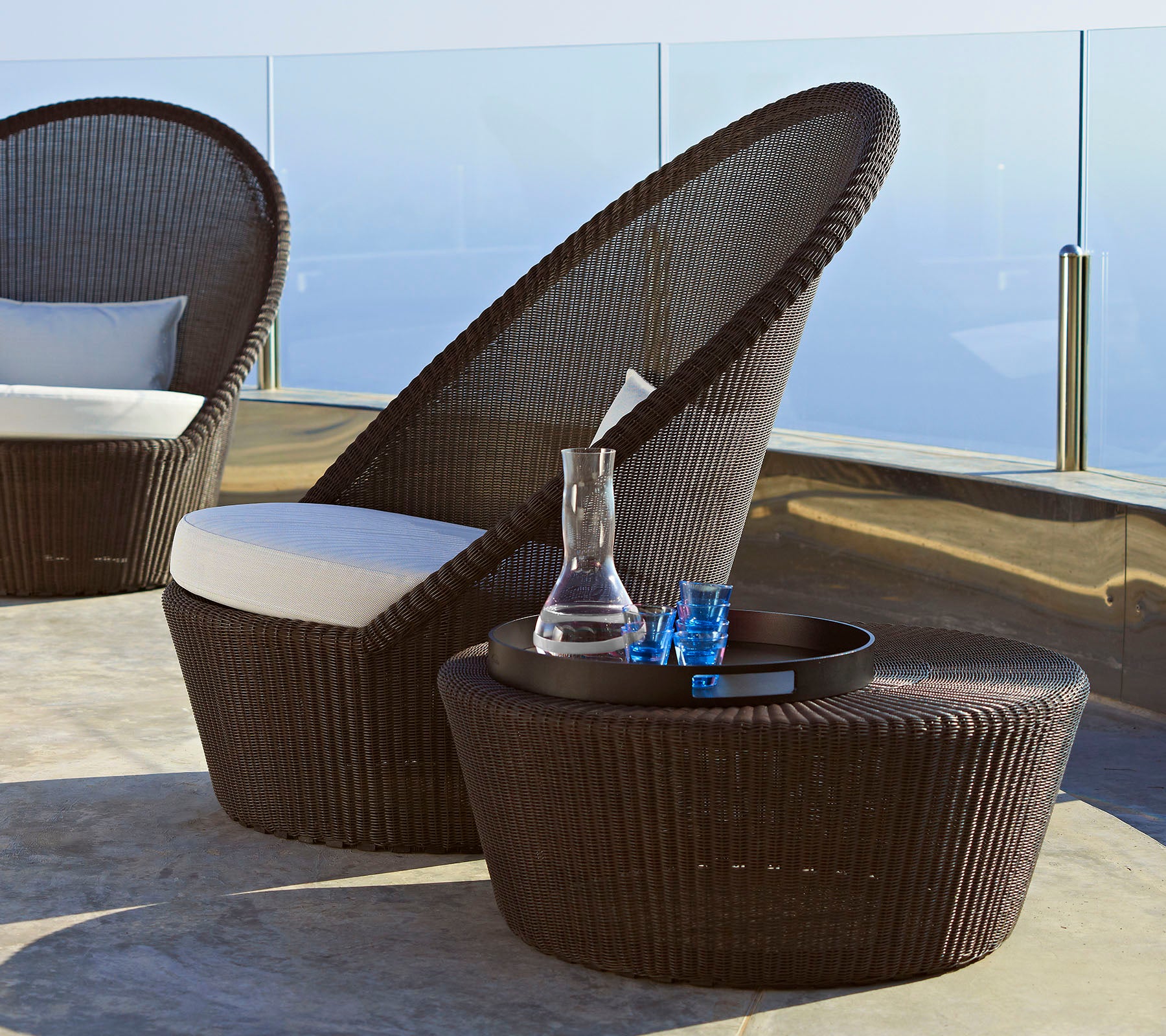 Silla de mimbre marrón elegante con respaldo alto y cojín blanco, acompañada de una mesa auxiliar a juego y artículos decorativos.