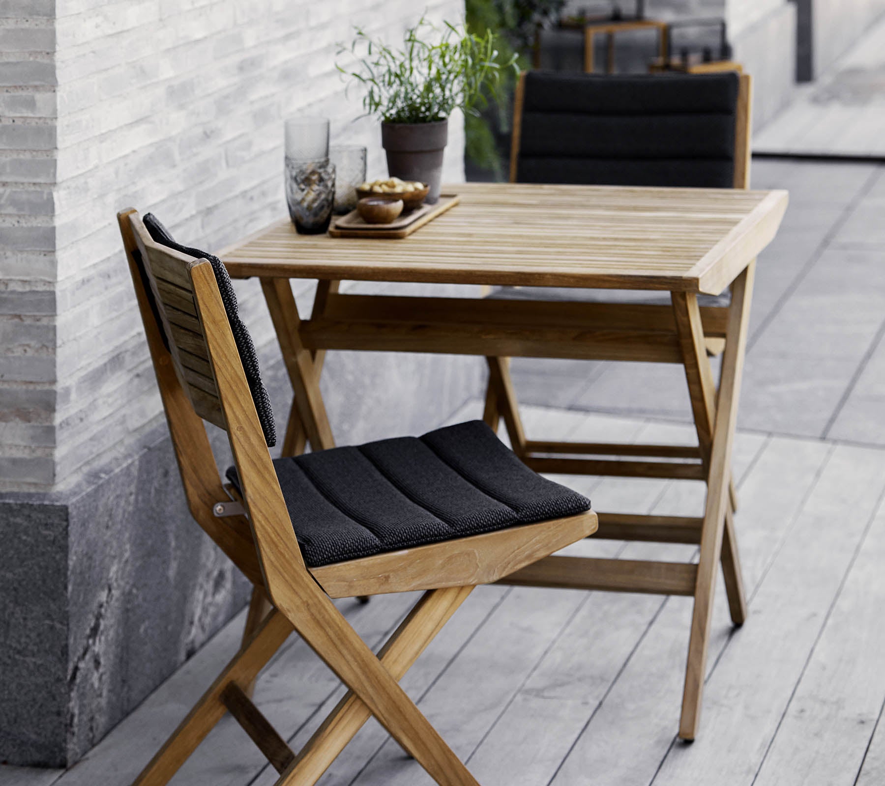 Conjunto de mesa y sillas de madera con cojines negros, adornado con plantas en macetas y cristalería decorativa. Perfecto para un ambiente acogedor.