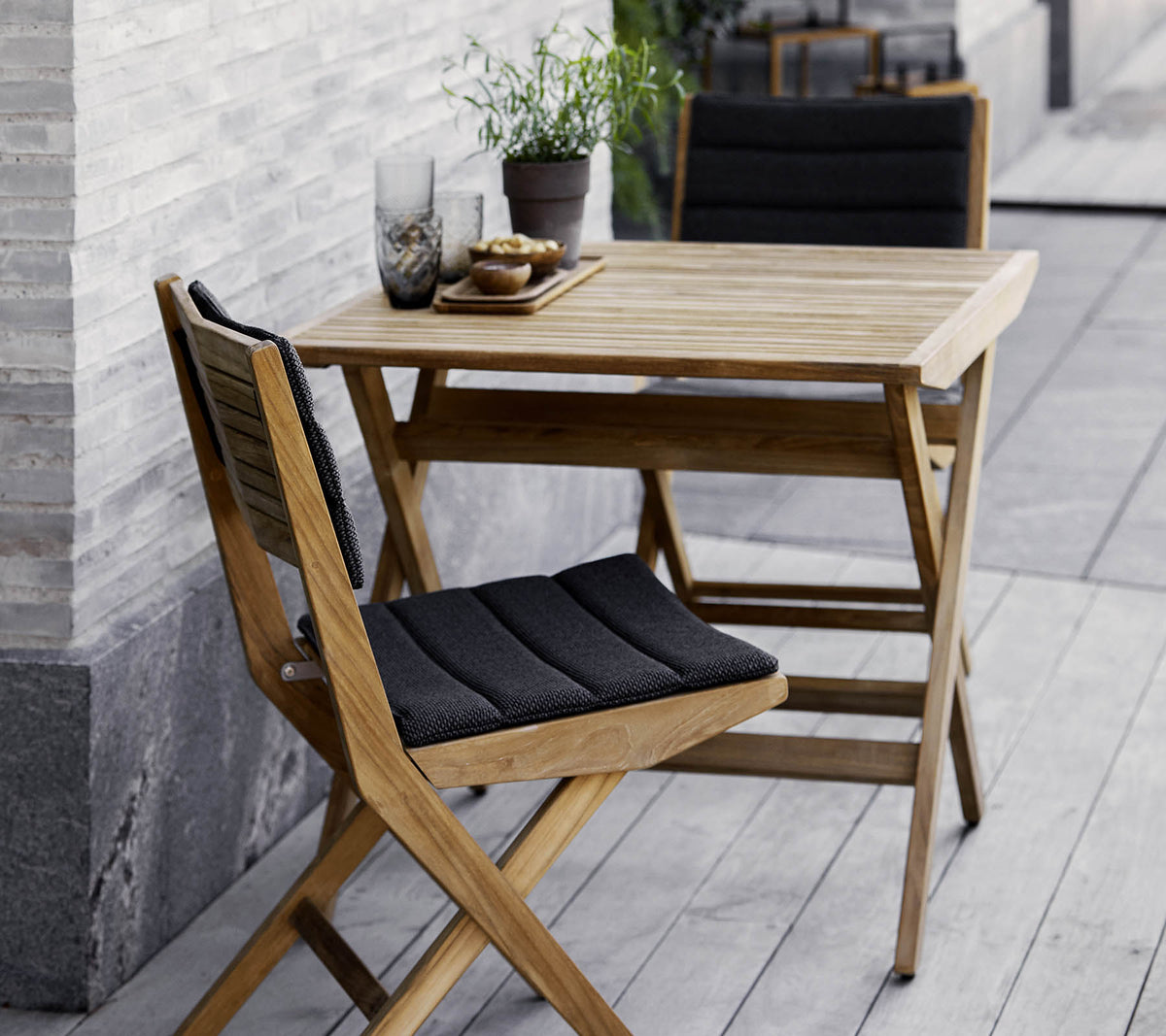 Conjunto de mesa y sillas de madera con cojines negros, adornado con plantas en macetas y cristalería decorativa. Perfecto para un ambiente acogedor.