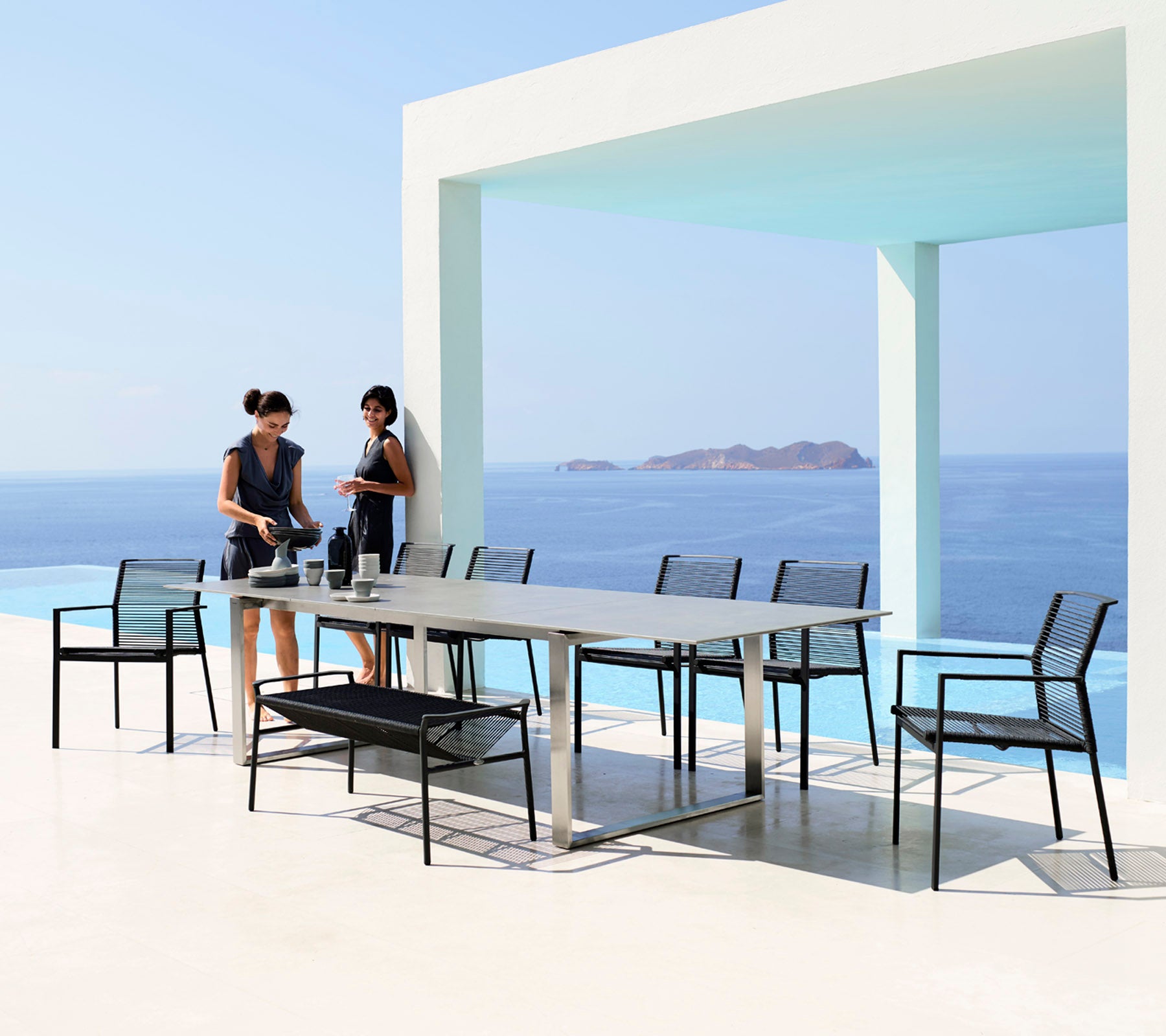 Dos mujeres preparan una mesa de comedor con elegantes sillas negras contra el telón de fondo de un sereno mar y cielo azul.