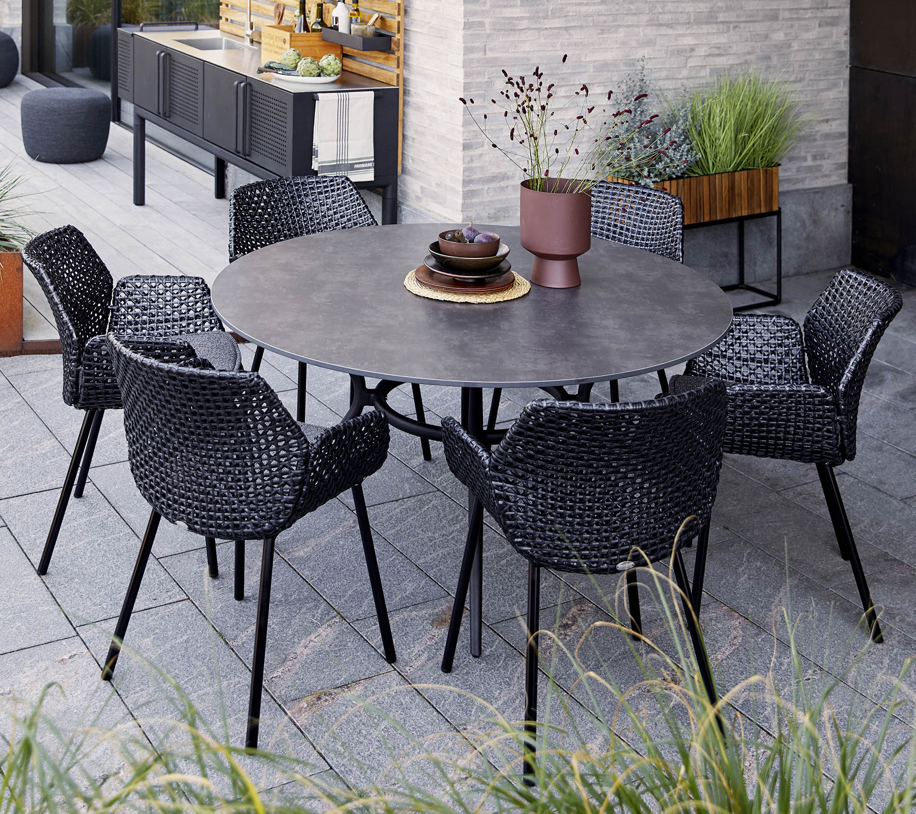 Mesa redonda con seis sillas negras tejidas, con un cuenco decorativo y una planta en maceta, situada entre la vegetación y una decoración moderna.