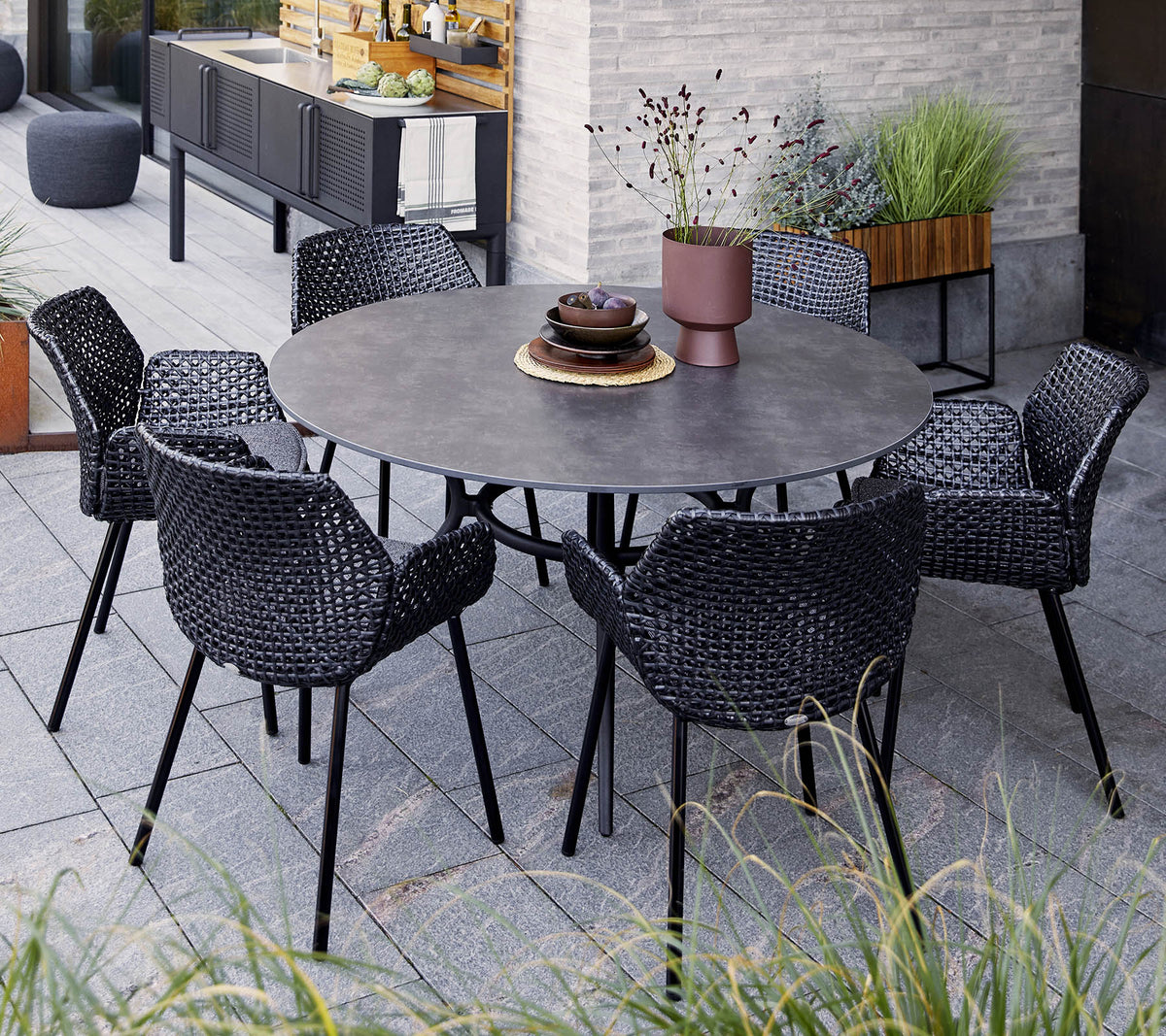 Mesa redonda con seis sillas negras tejidas, con un cuenco decorativo y una planta en maceta, situada entre la vegetación y una decoración moderna.