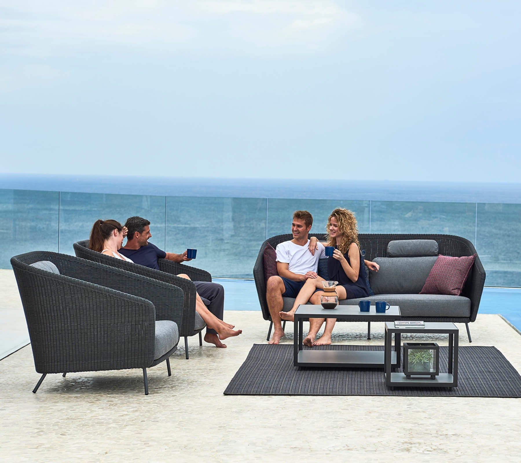 Un grupo de cuatro personas socializando en muebles de exterior modernos con vista al océano, disfrutando de bebidas y conversación.