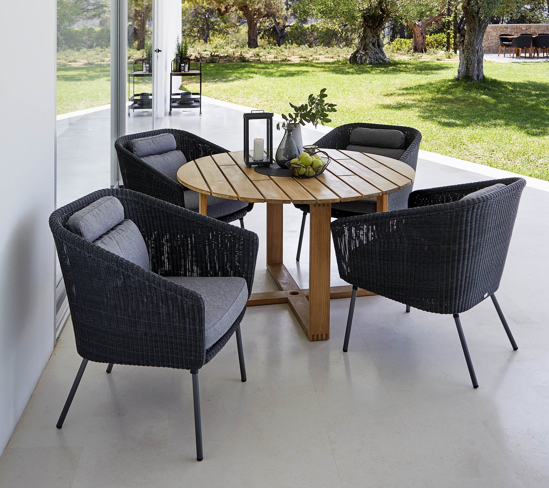 Una mesa redonda de madera rodeada por cuatro sillas trenzadas negras y elegantes con cojines grises, acentuada por una linterna decorativa y vegetación.