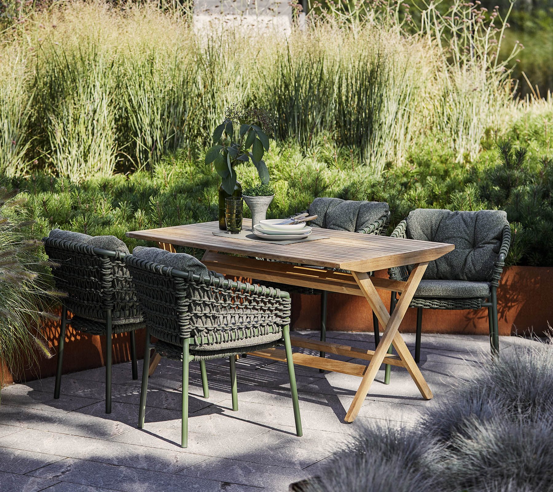 Un conjunto de comedor al aire libre elegante que presenta una mesa de madera y cuatro sillas trenzadas, rodeado de vegetación exuberante y plantas decorativas.