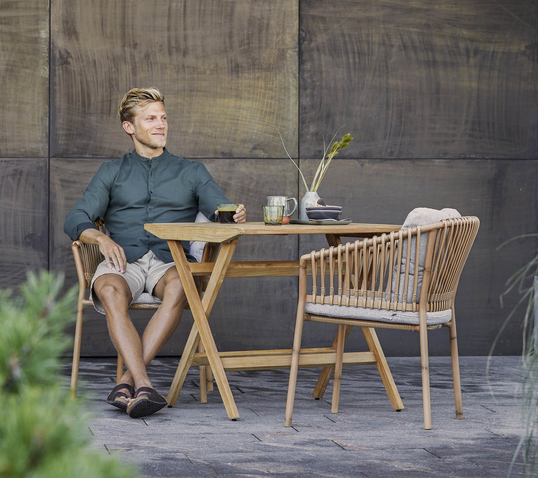 Un hombre está sentado relajado en una elegante mesa de madera con una bebida ligera, rodeado de vegetación y un fondo texturizado.