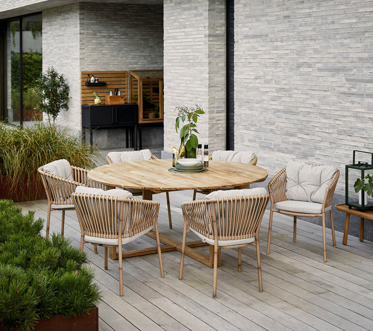 Mesa de comedor de madera redonda rodeada de sillas tapizadas, con un centro de mesa decorativo de planta, contra una pared de piedra texturizada.