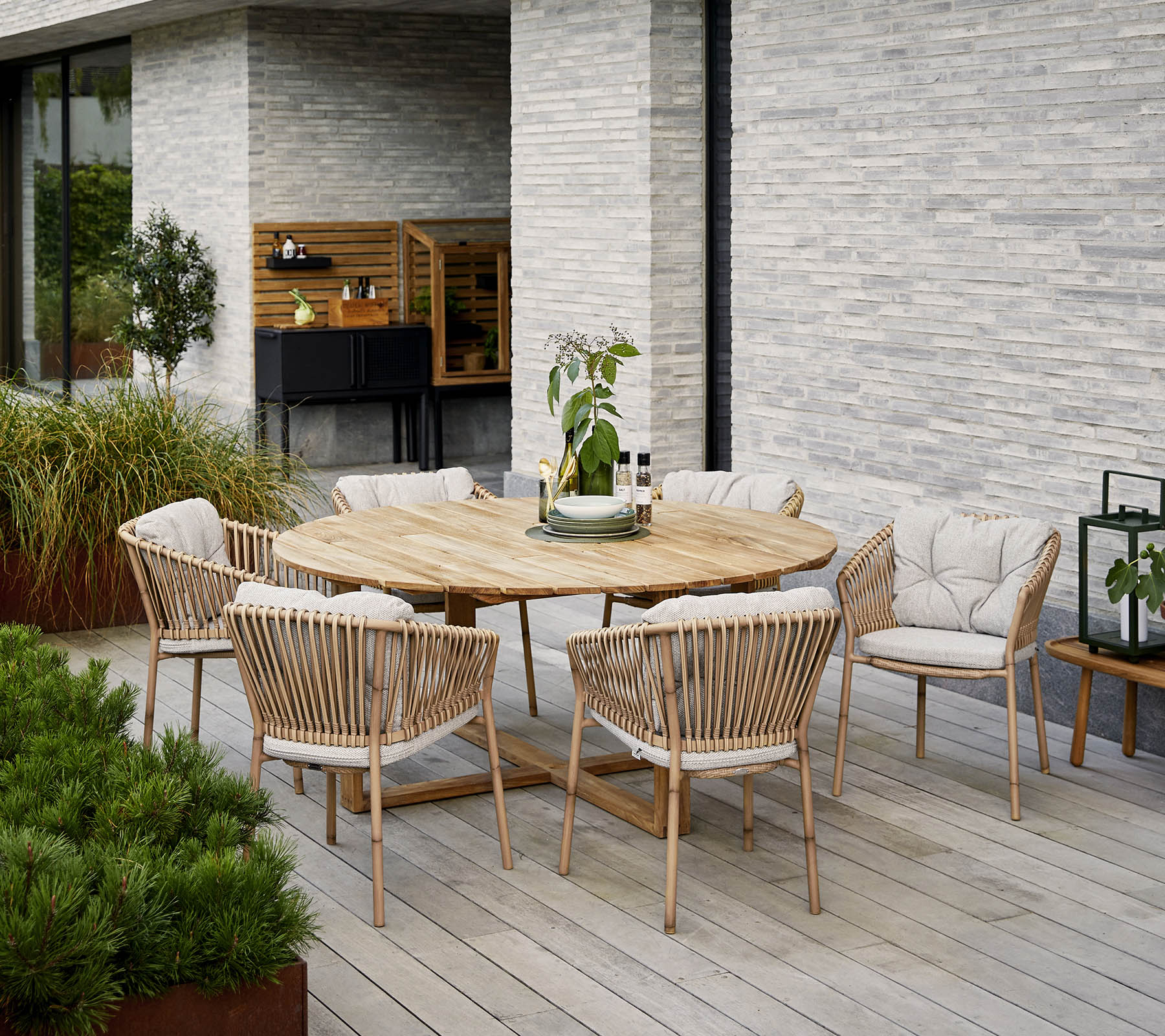 Mesa de comedor de madera redonda rodeada de sillas tapizadas, con un centro de mesa decorativo de planta, contra una pared de piedra texturizada.