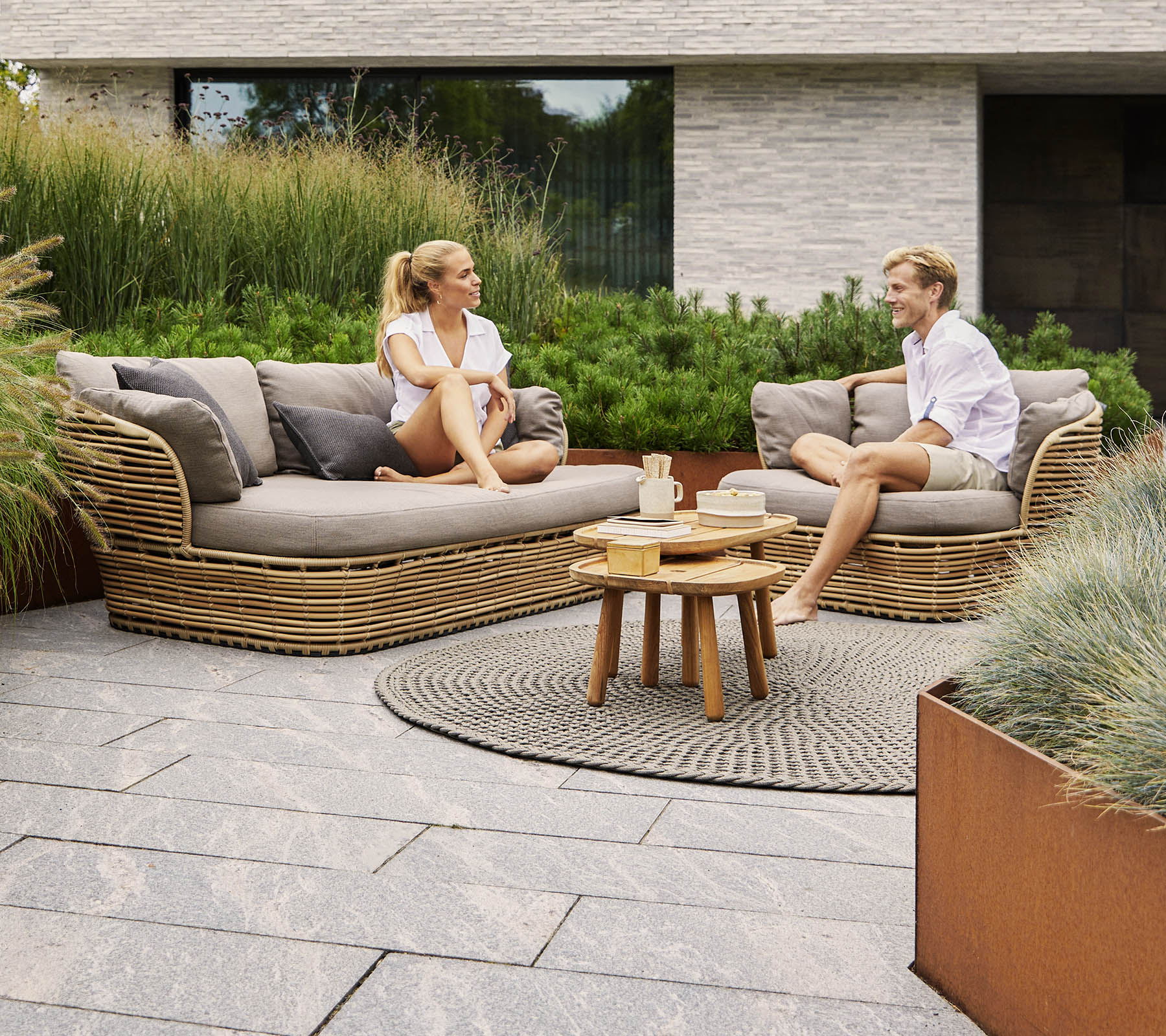 Dos personas conversan mientras están sentadas en un sofá curvado y elegante, rodeadas de vegetación y decoración moderna, con una pequeña mesa de madera entre ellas.