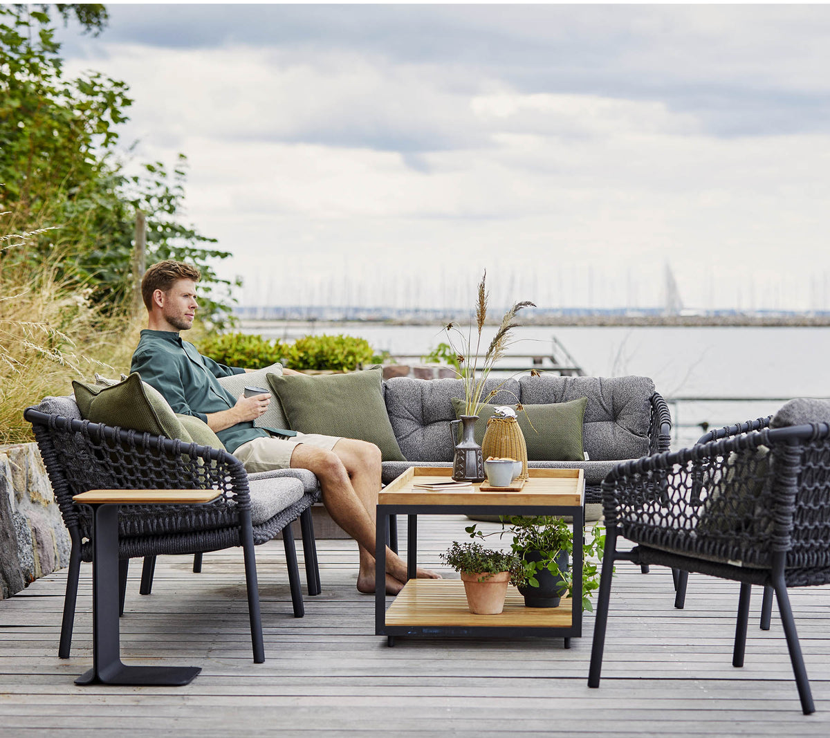 Una zona de estar cómoda con un hombre relajándose, con sillas de mimbre elegantes y una mesa de café de madera adornada con plantas y objetos decorativos.