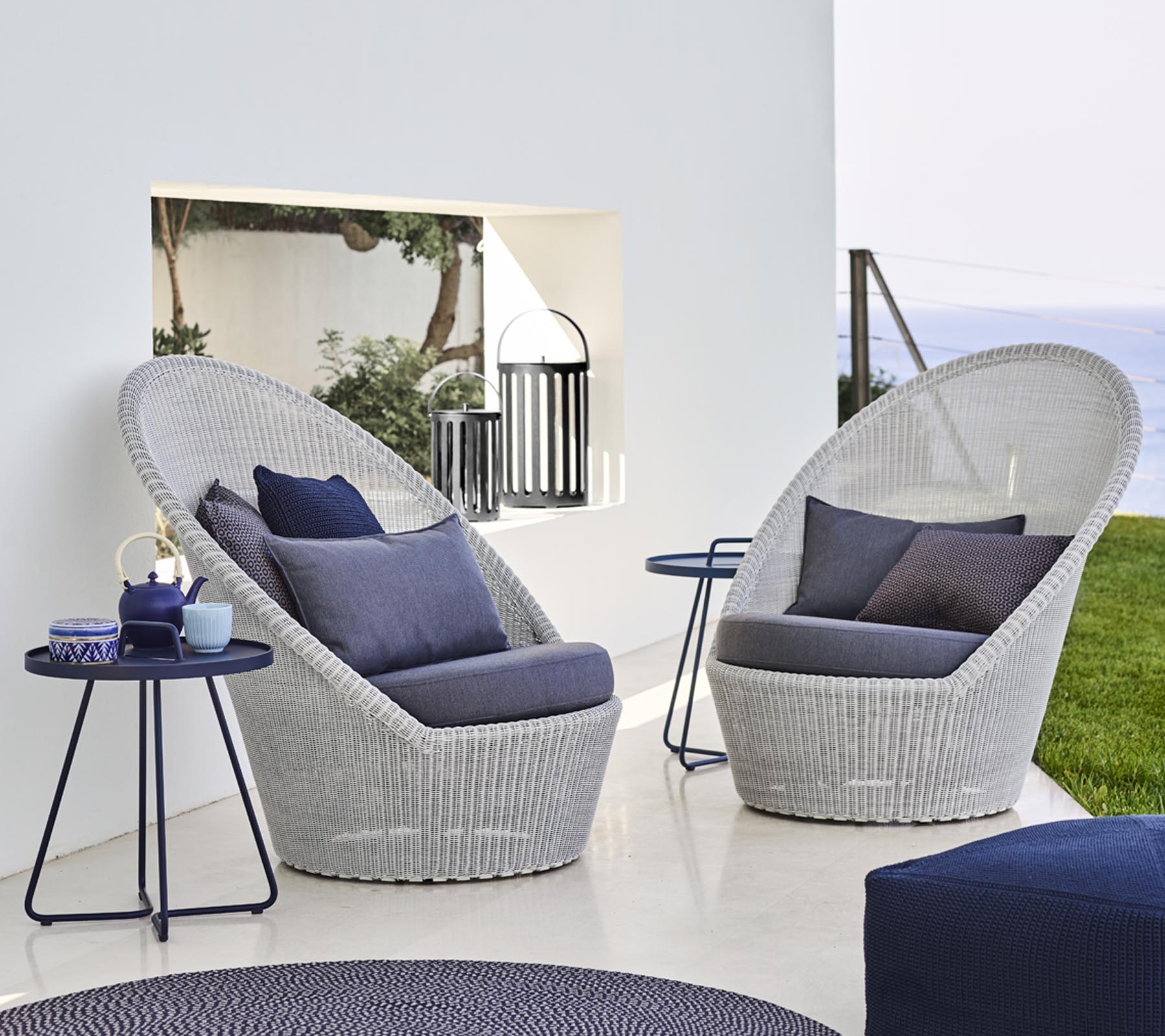 Dos sillones de mimbre elegantes con cojines azules al lado de una mesa pequeña, situados en un fondo sereno.