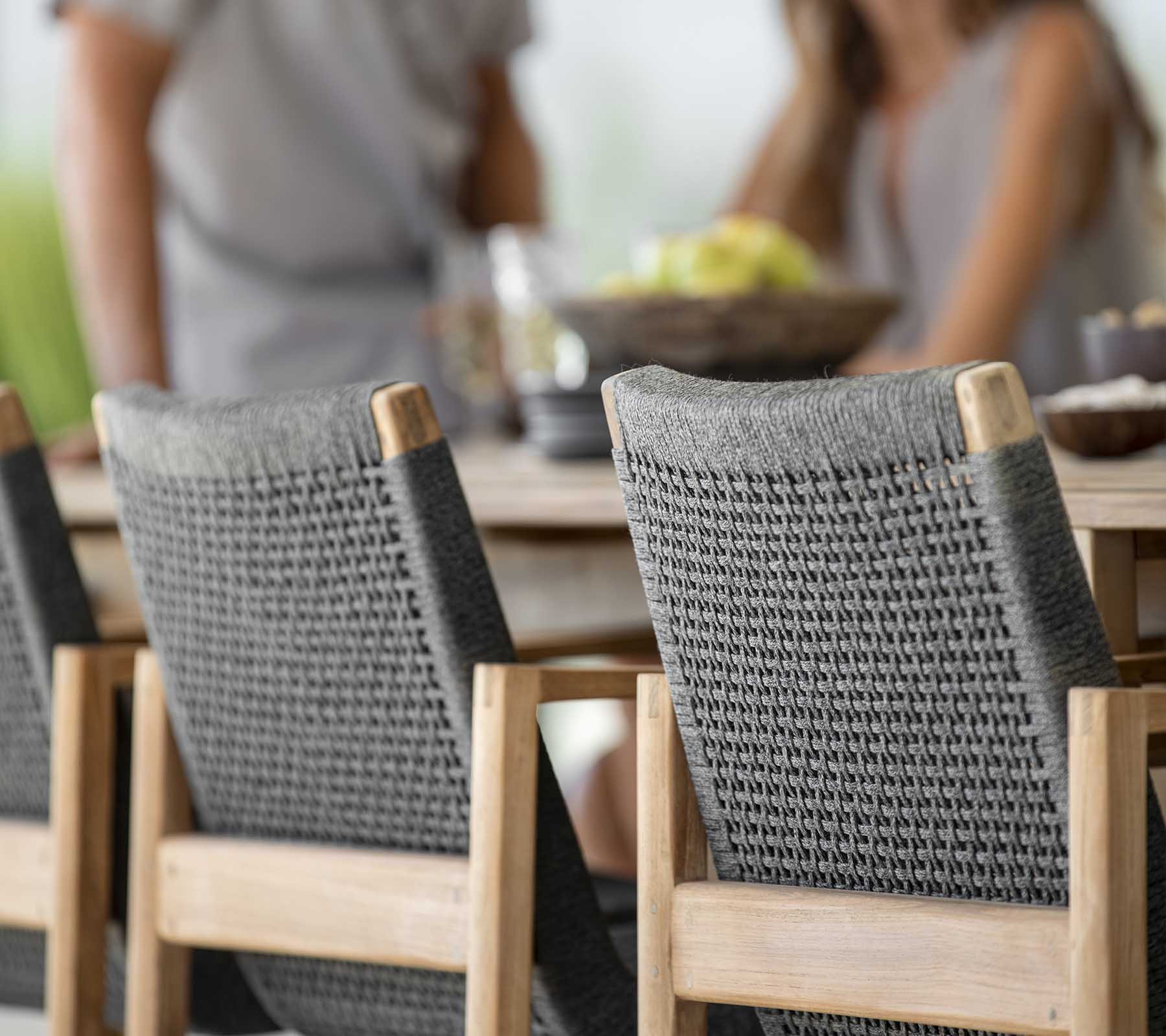 Escena de comedor al aire libre con una mesa de madera y dos sillas trenzadas grises elegantes, donde la gente disfruta de una comida junta.