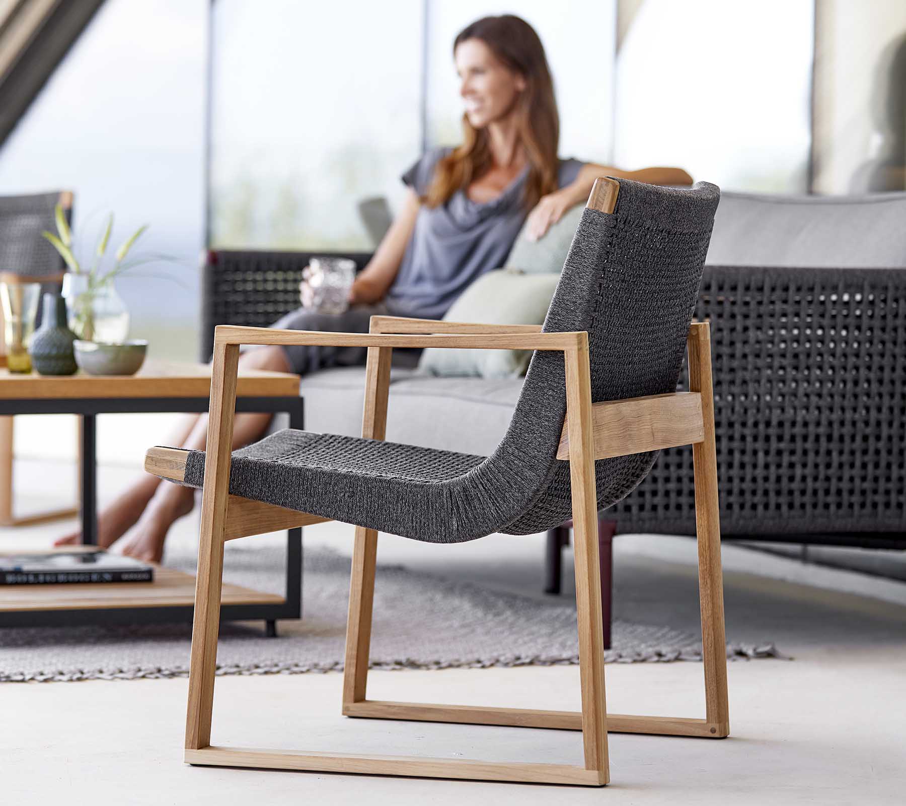Una silla moderna con un elegante marco de madera y tela tejida, situada junto a una mujer que se relaja y disfruta de una bebida.
