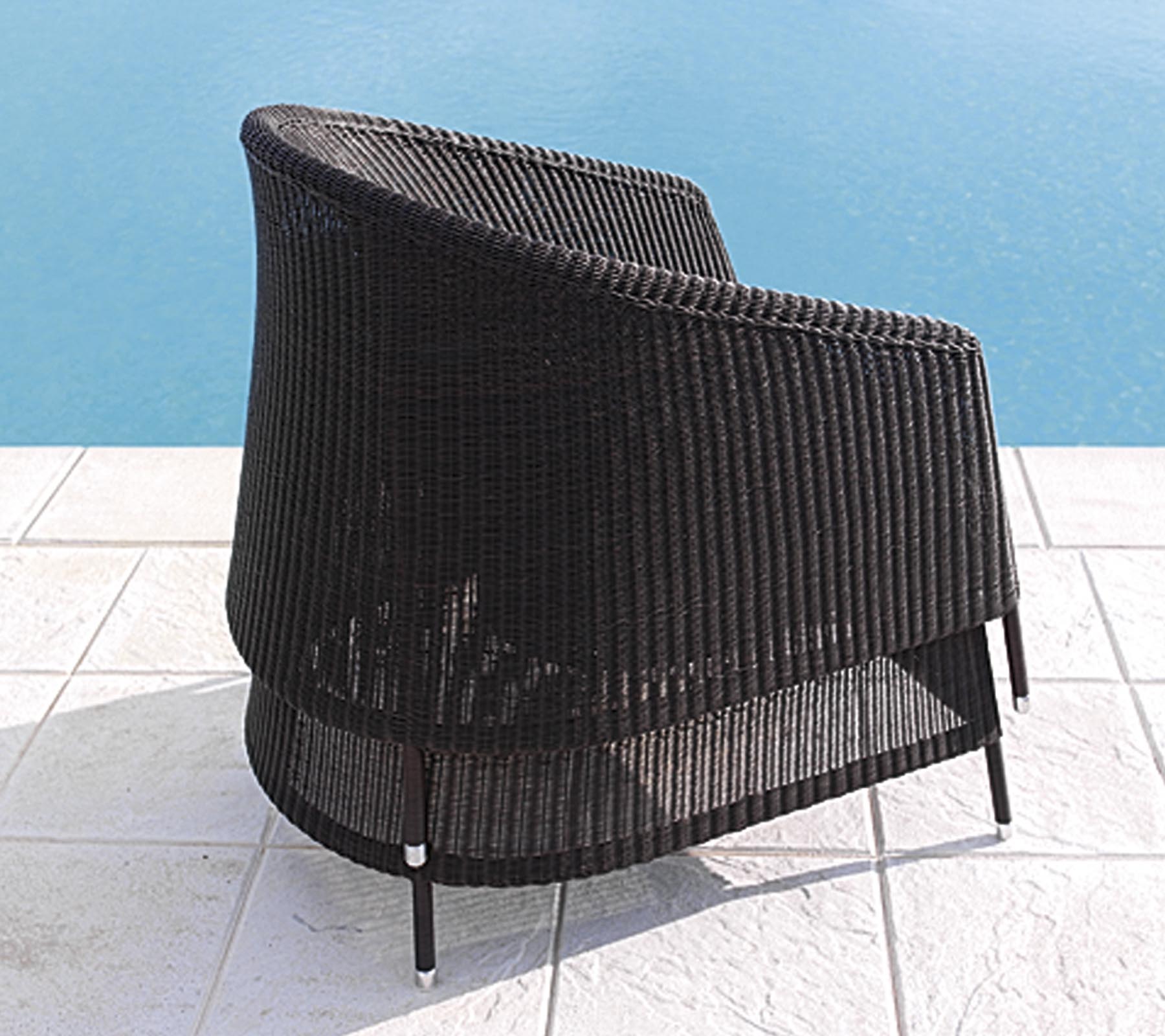 Silla de ratán negro curvada con un diseño elegante, situada cerca de una piscina en un suelo de baldosas blancas.