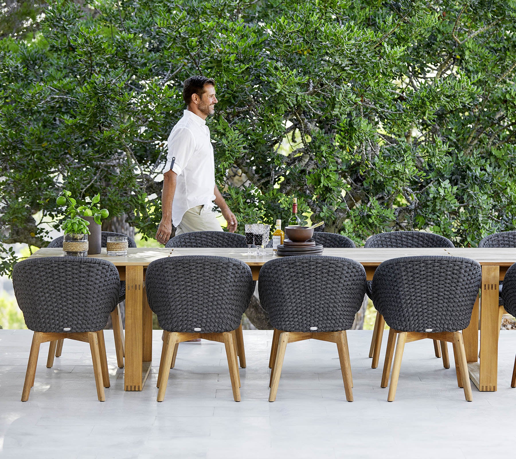 Un hombre pasa junto a una gran mesa de comedor de madera rodeada de sillas modernas de mimbre y vegetación al fondo.