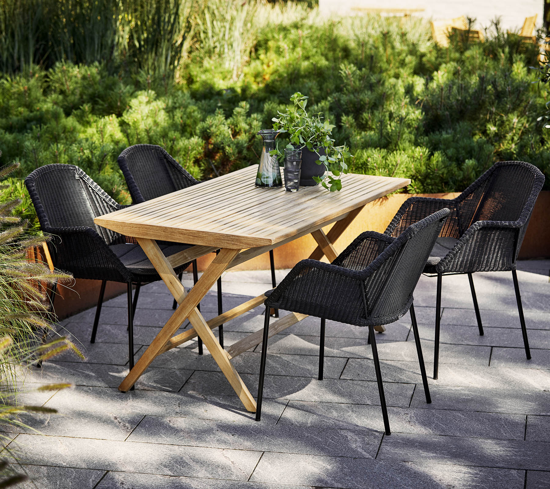 Una mesa de comedor de madera rodeada de cuatro sillas trenzadas negras, con vegetación en el fondo y artículos decorativos en la mesa.