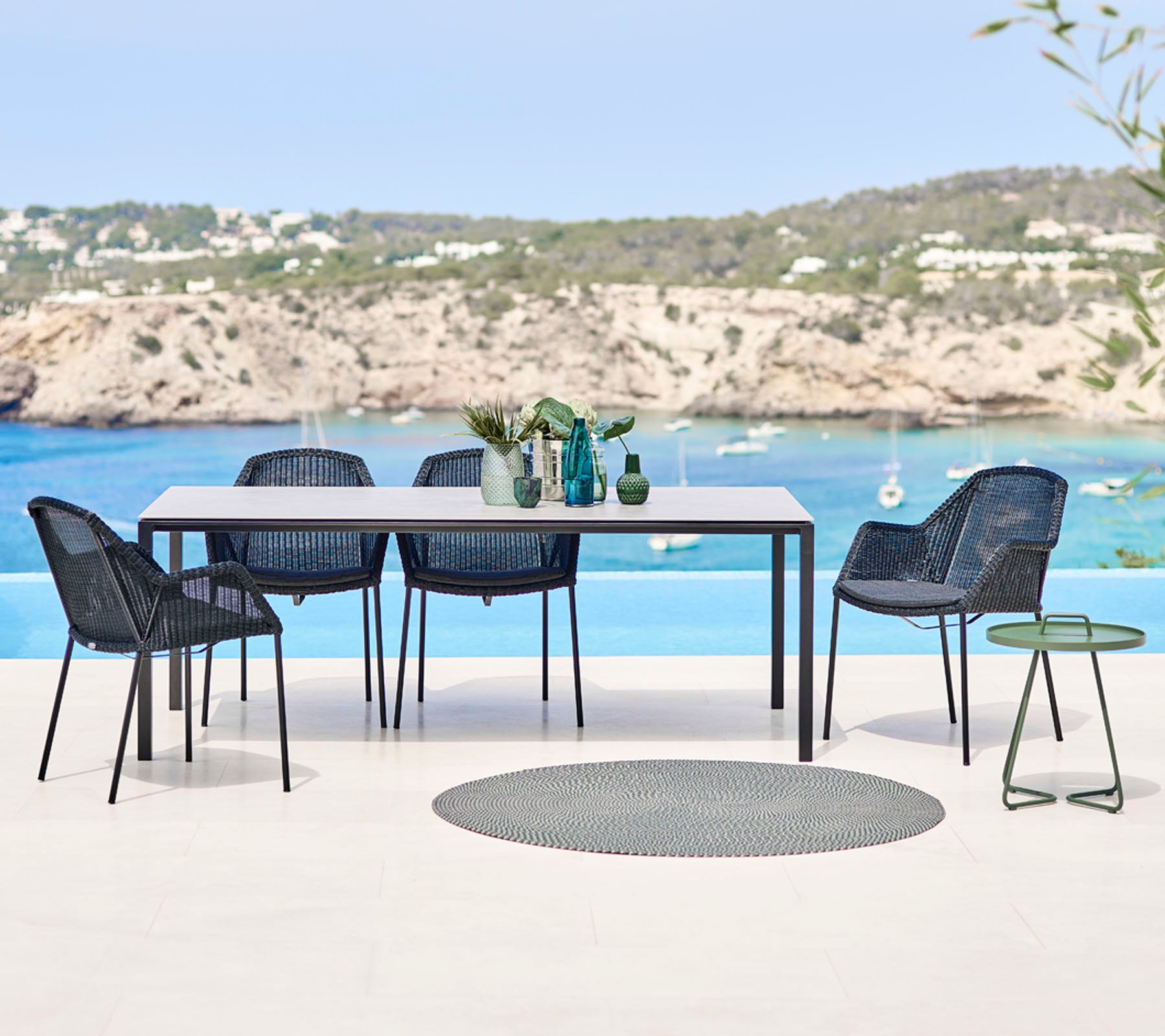 Un juego de comedor al aire libre moderno que incluye una mesa rectangular, sillas trenzadas y una mesa auxiliar pequeña, con un fondo de playa pintoresco.