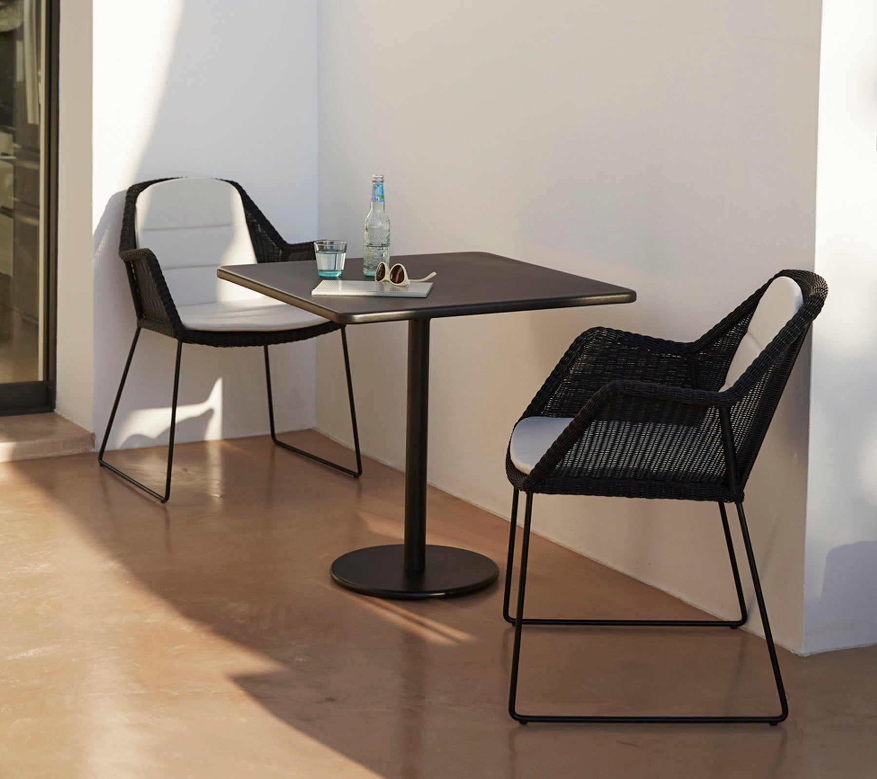Mesa moderna con dos elegantes sillas negras, acompañada de una botella de agua y gafas de sol, situada contra un fondo minimalista.