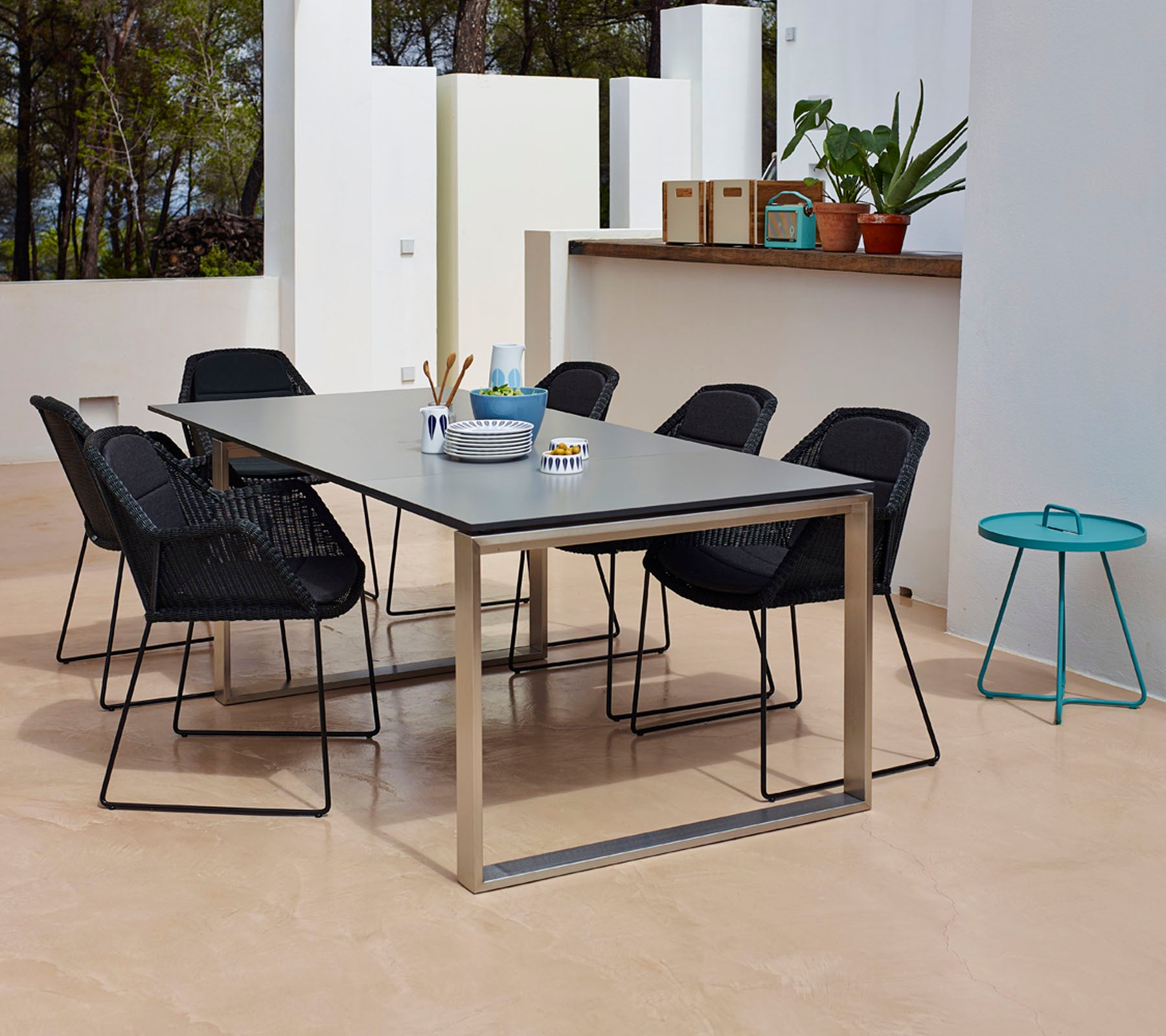 Un conjunto de comedor moderno con una mesa rectangular de superficie lisa, rodeada de seis sillas negras trenzadas y una pequeña mesa lateral color verde azulado.