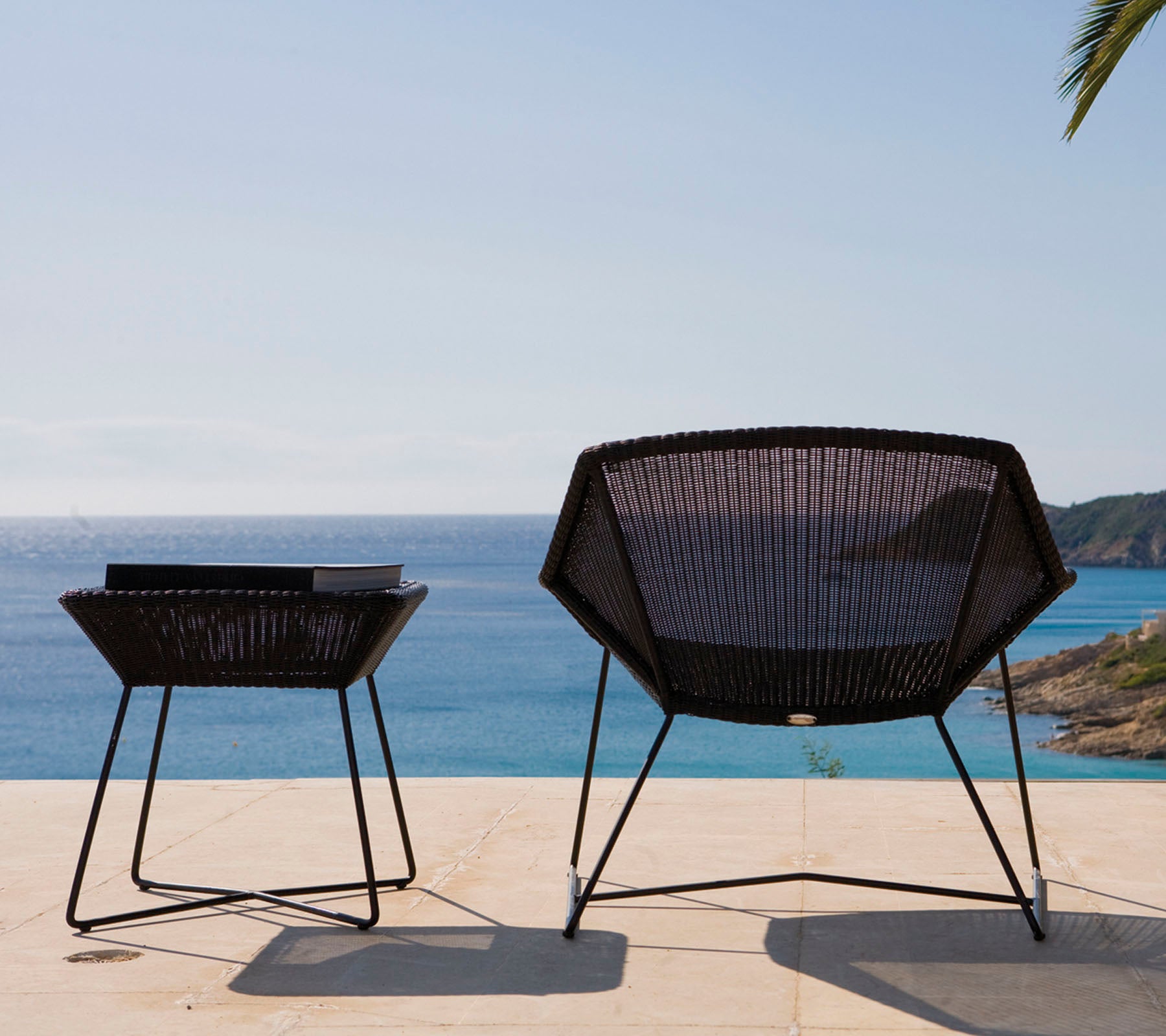 Dos sillas de exterior modernas y negras con un diseño geométrico están situadas junto a una mesa, con vista a un mar tranquilo bajo un cielo despejado.