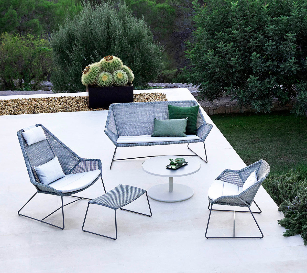 Elegante disposición de asientos al aire libre que incluye un sofá, dos sillones, un reposapiés y una mesa de café, complementada con vegetación en el fondo.