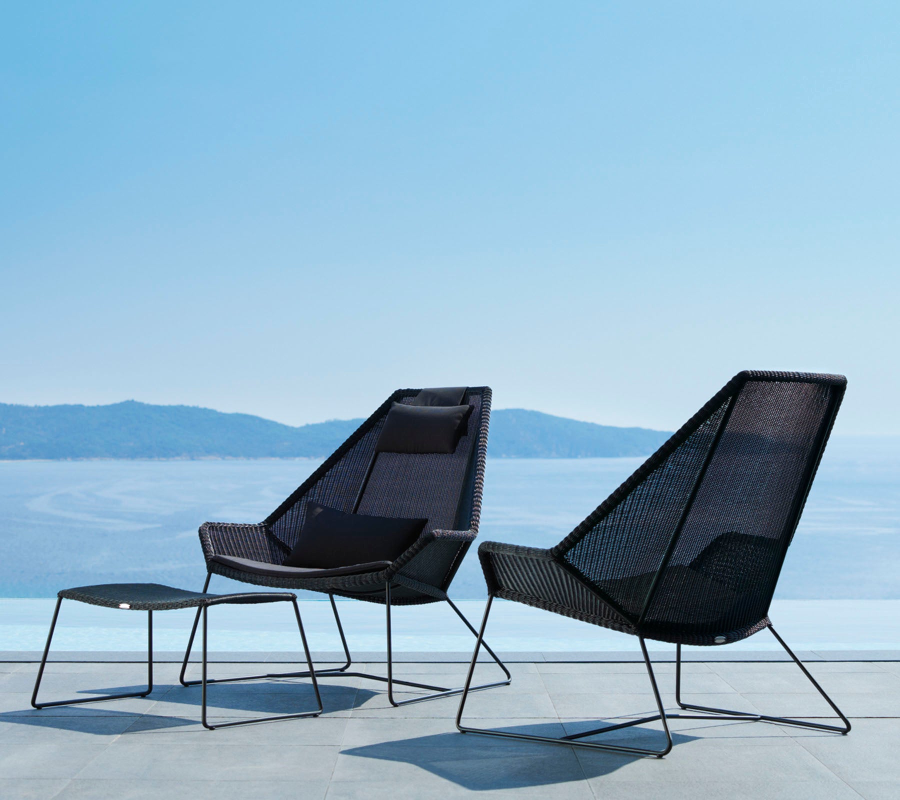 Dos sillas modernas y elegantes con líneas limpias y un diseño de malla abierta, acompañadas de un reposapiés a juego, con vistas a una serena vista del agua.