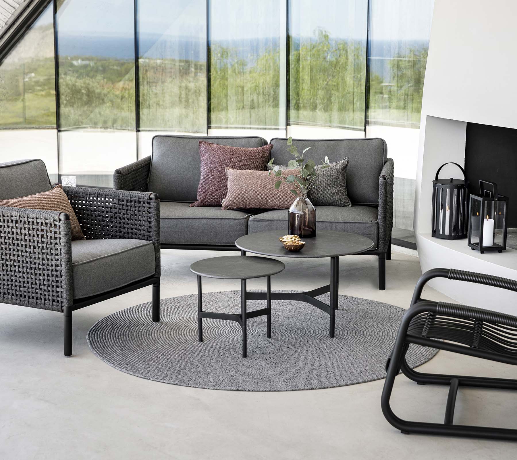 Área de estar elegante con un sofá gris con cojines, mesa de café redonda y sillas decorativas sobre una alfombra circular. La luz natural realza la atmósfera.