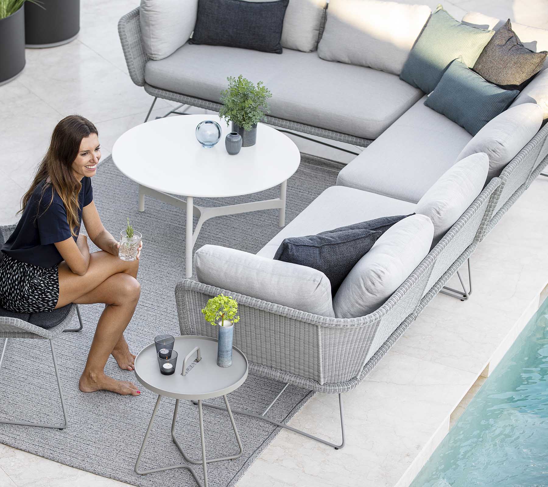 Un elegante arreglo de asientos al aire libre con un sofá, cojines decorativos y una mesa pequeña, complementado por plantas y una piscina cercana.