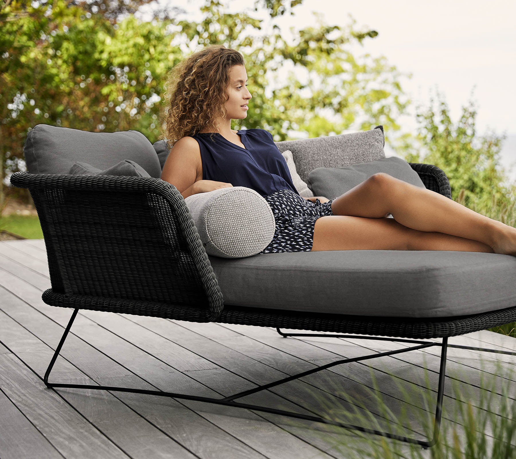 Una mujer se relaja en una silla lounge elegante y acolchada adornada con cojines, rodeada de vegetación.