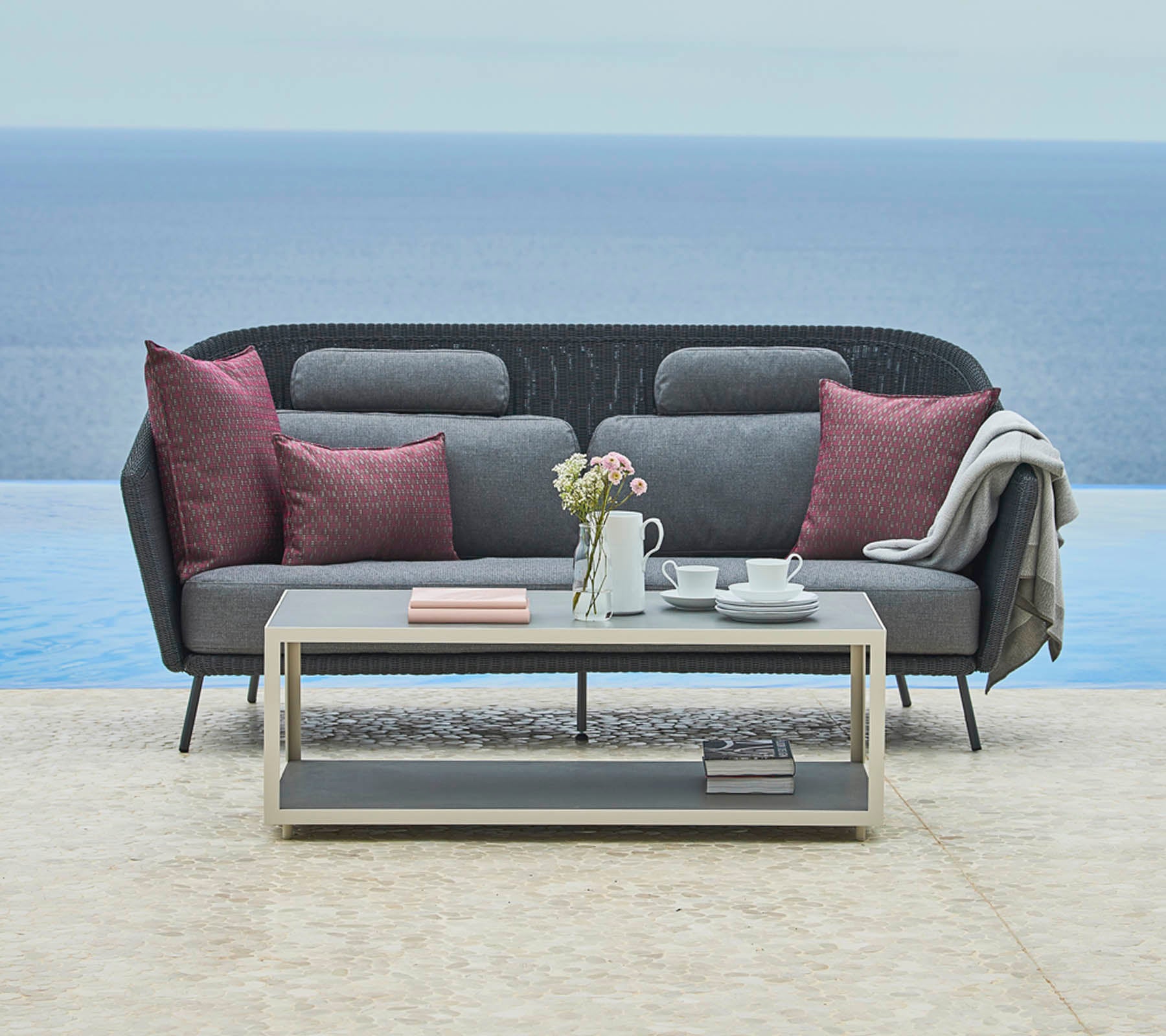 Sofa gris elegante con cojines decorativos y una manta acogedora, complementado por una mesa de café elegante con una tetera y libros, situado frente a un paisaje sereno.