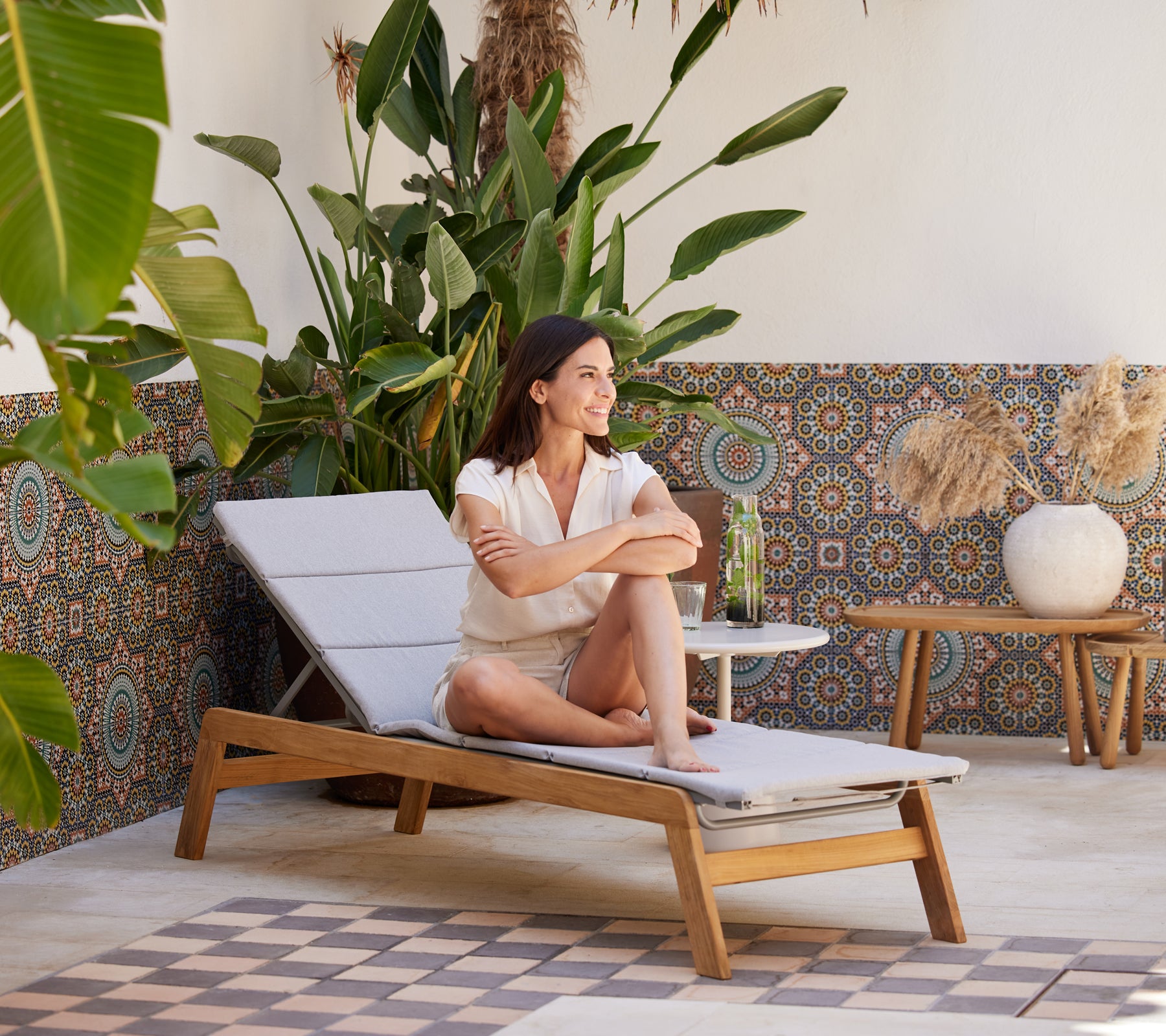 Una mujer se relaja en una chaise longue rodeada de plantas vibrantes y decoraciones con patrones, disfrutando de una bebida refrescante.