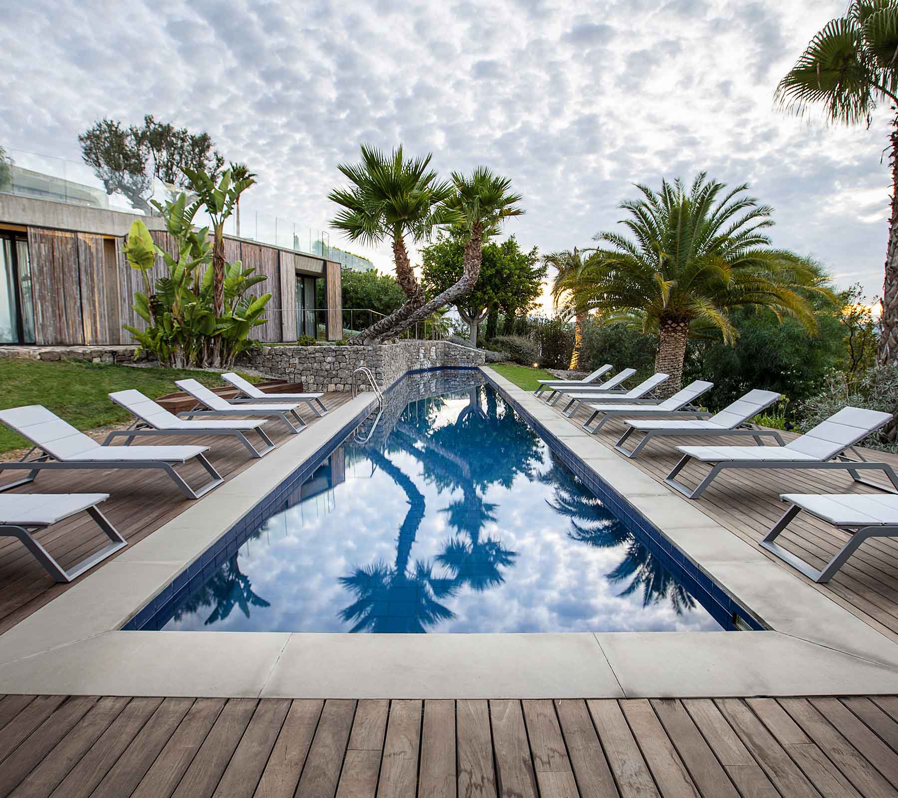 Escena serena con una piscina rodeada de tumbonas y palmeras, que refleja el cielo y la exuberante vegetación.