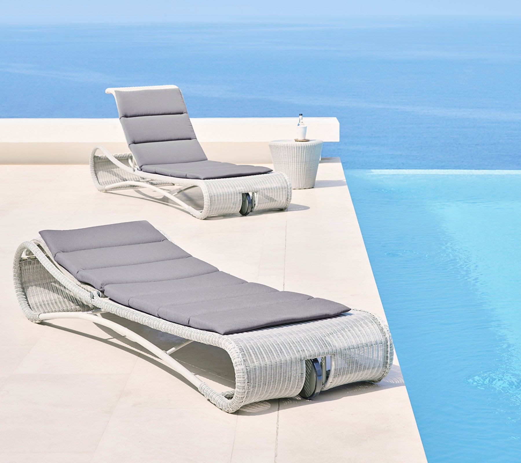 Dos elegantes sillones con cojines grises al lado de una serena piscina infinita azul, creando un espacio exterior relajante.