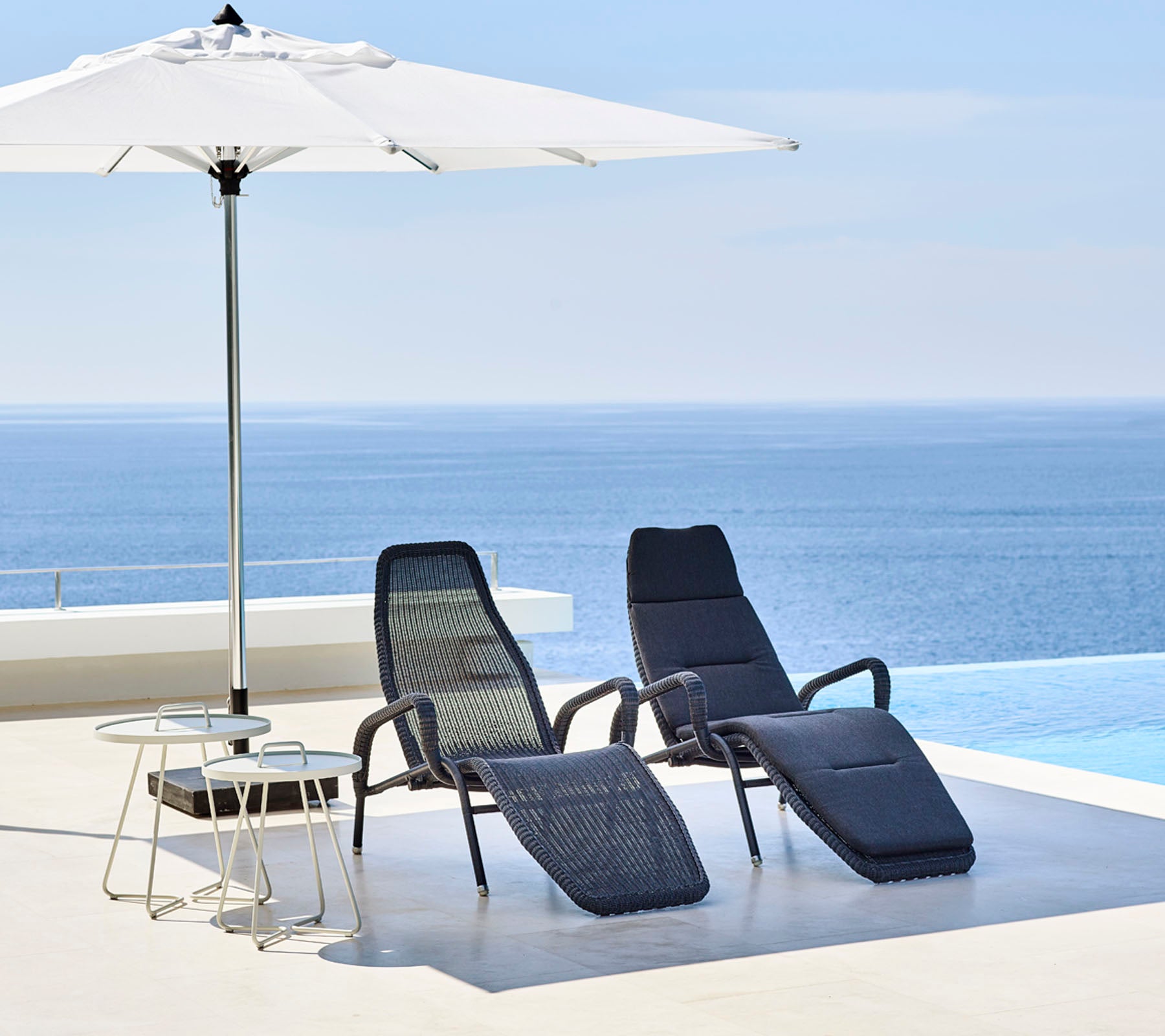Dos sillas de lounge elegantes y una mesa pequeña están bajo una gran sombrilla, con un sereno mar azul de fondo.