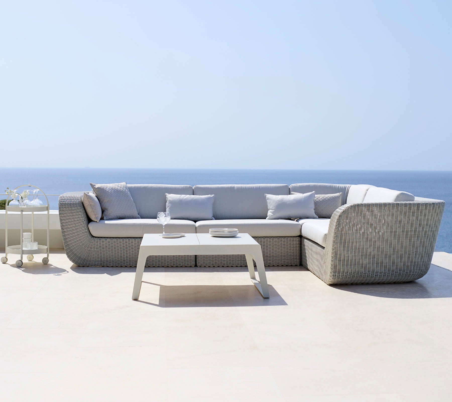Sofá seccional moderno con cojines gris claro combinado con una mesa de café blanca y elegante, situada frente a un sereno paisaje oceánico.