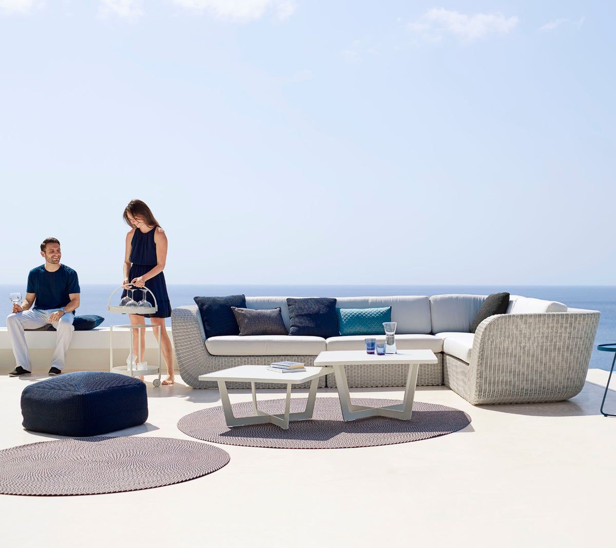 Un área de descanso al aire libre moderna con un sofá seccional, una mesa de café y alfombras decorativas, con dos personas disfrutando del espacio.
