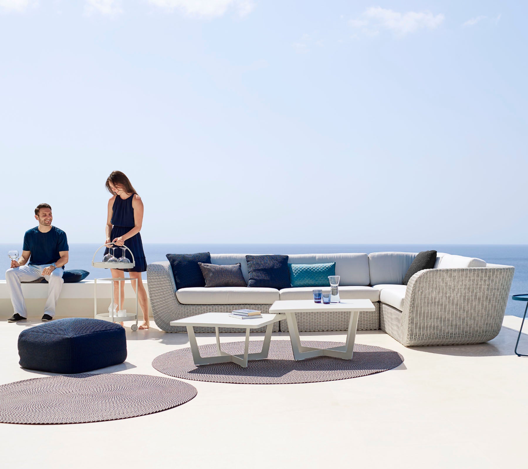 Un área de descanso al aire libre moderna con un sofá seccional, una mesa de café y alfombras decorativas, con dos personas disfrutando del espacio.