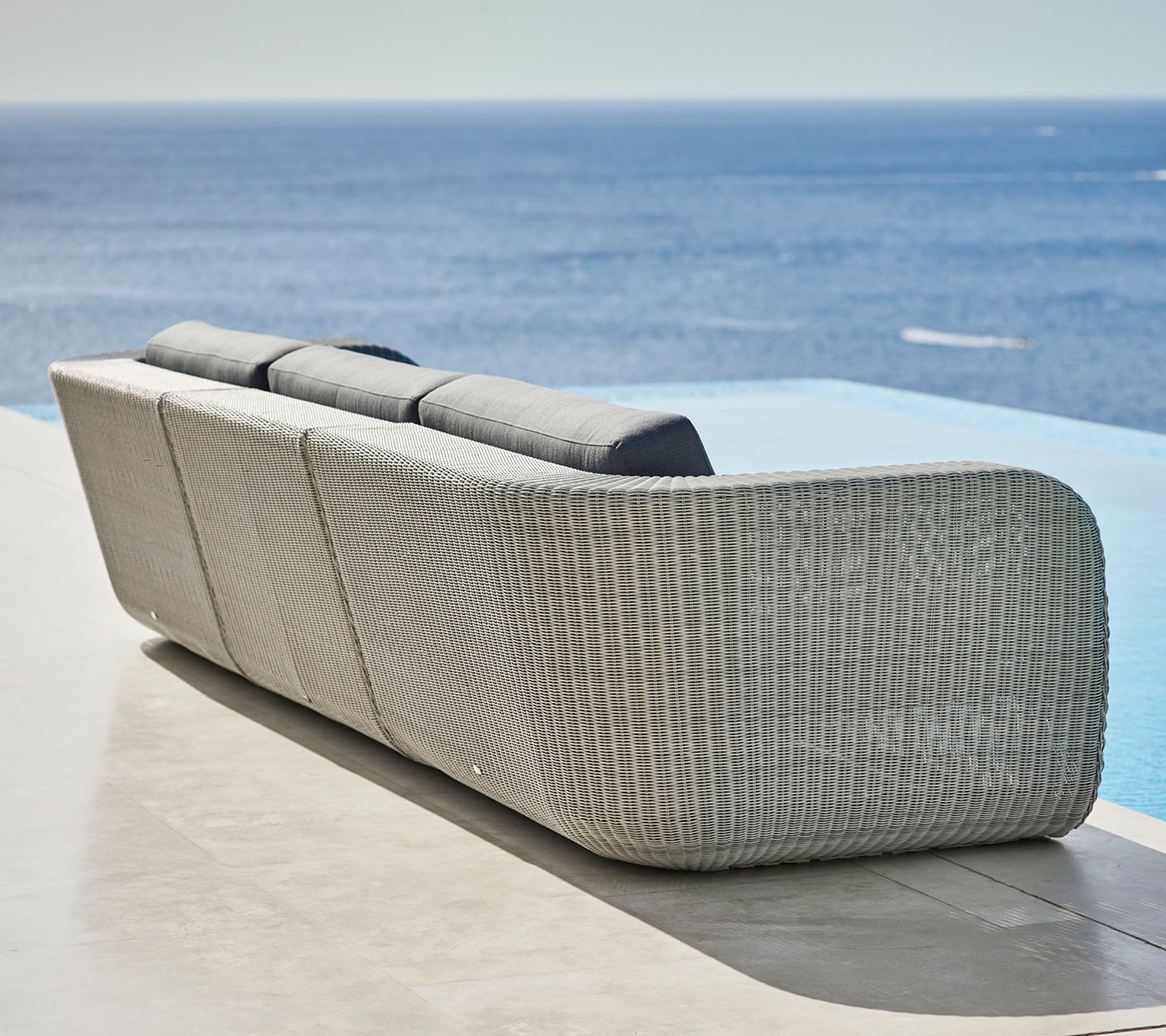 Sofá tejido con estilo, de diseño curvado y cojines mullidos, con vistas a un tranquilo océano. Ideal para relajarse y entornos al aire libre.