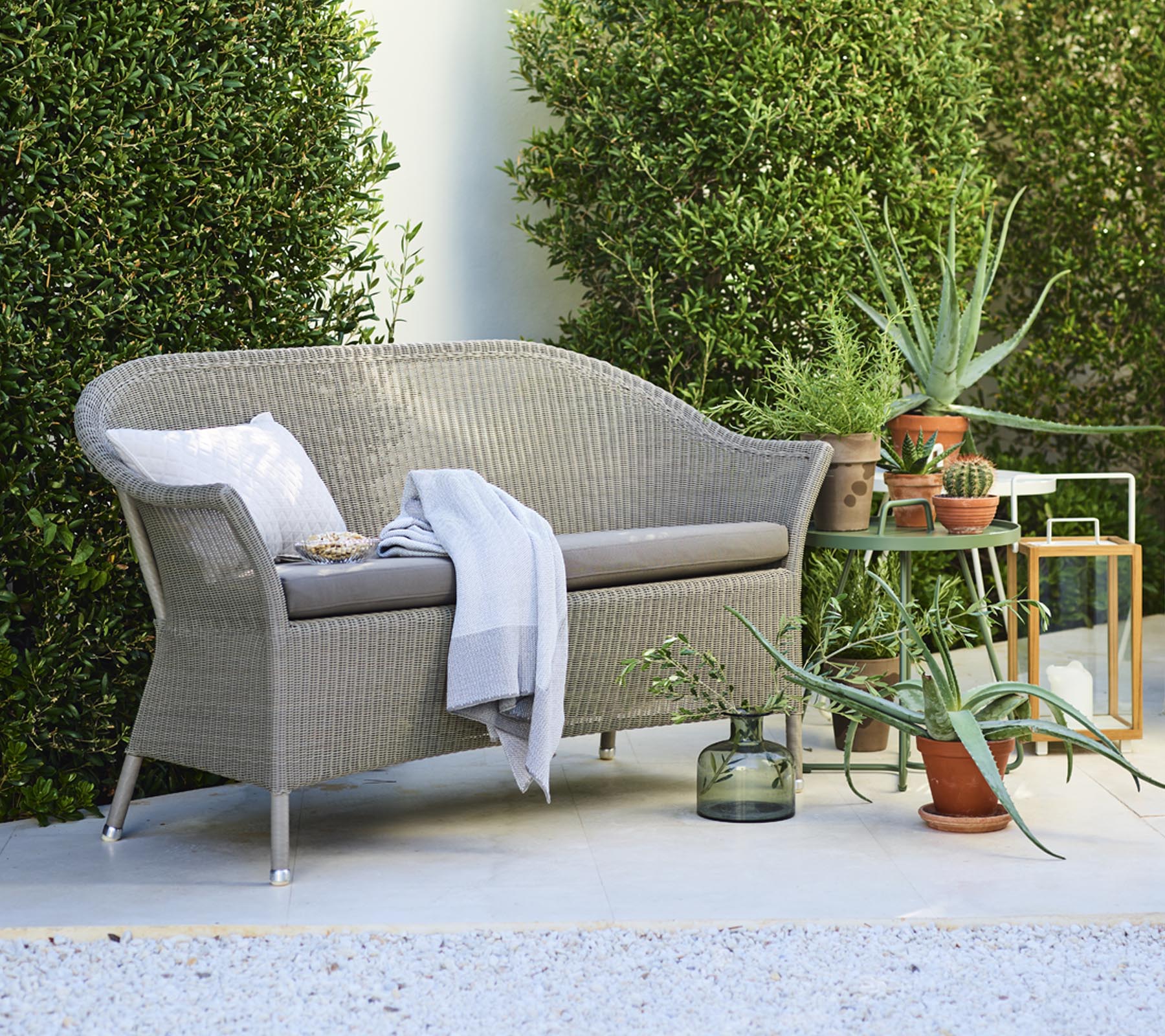 Un acogedor sofá loveseat con cojines decorativos, acompañado de plantas en macetas y una mesa auxiliar, rodeado de vegetación.