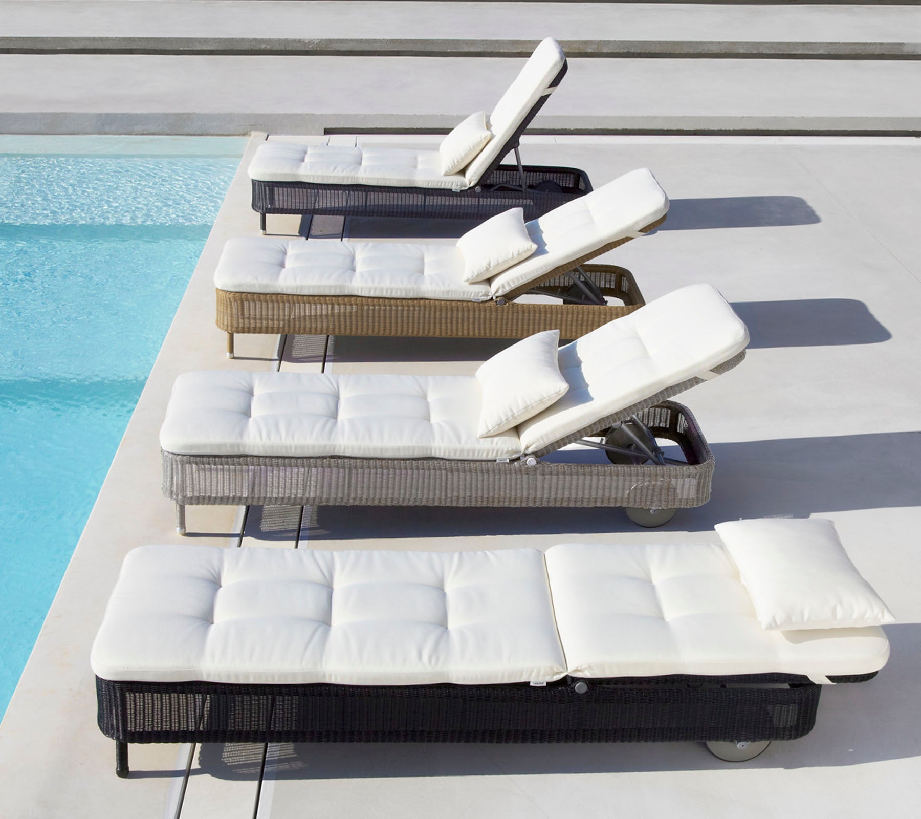 Cuatro tumbonas de varios colores con cojines blancos, situadas junto a una piscina, cada una con respaldo ajustable.