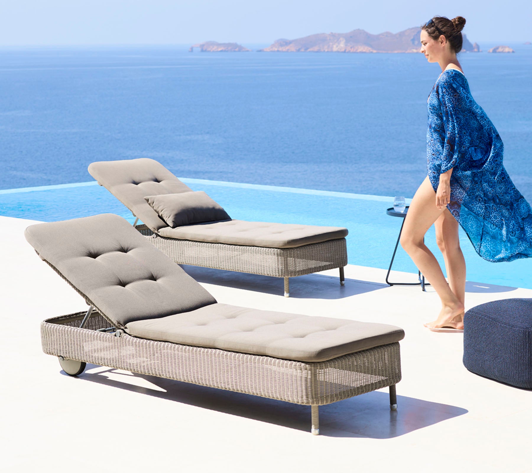 Una mujer con un cubre vestido azul fluido camina junto a dos elegantes sillas reclinables junto a una piscina, con vista a un sereno océano.
