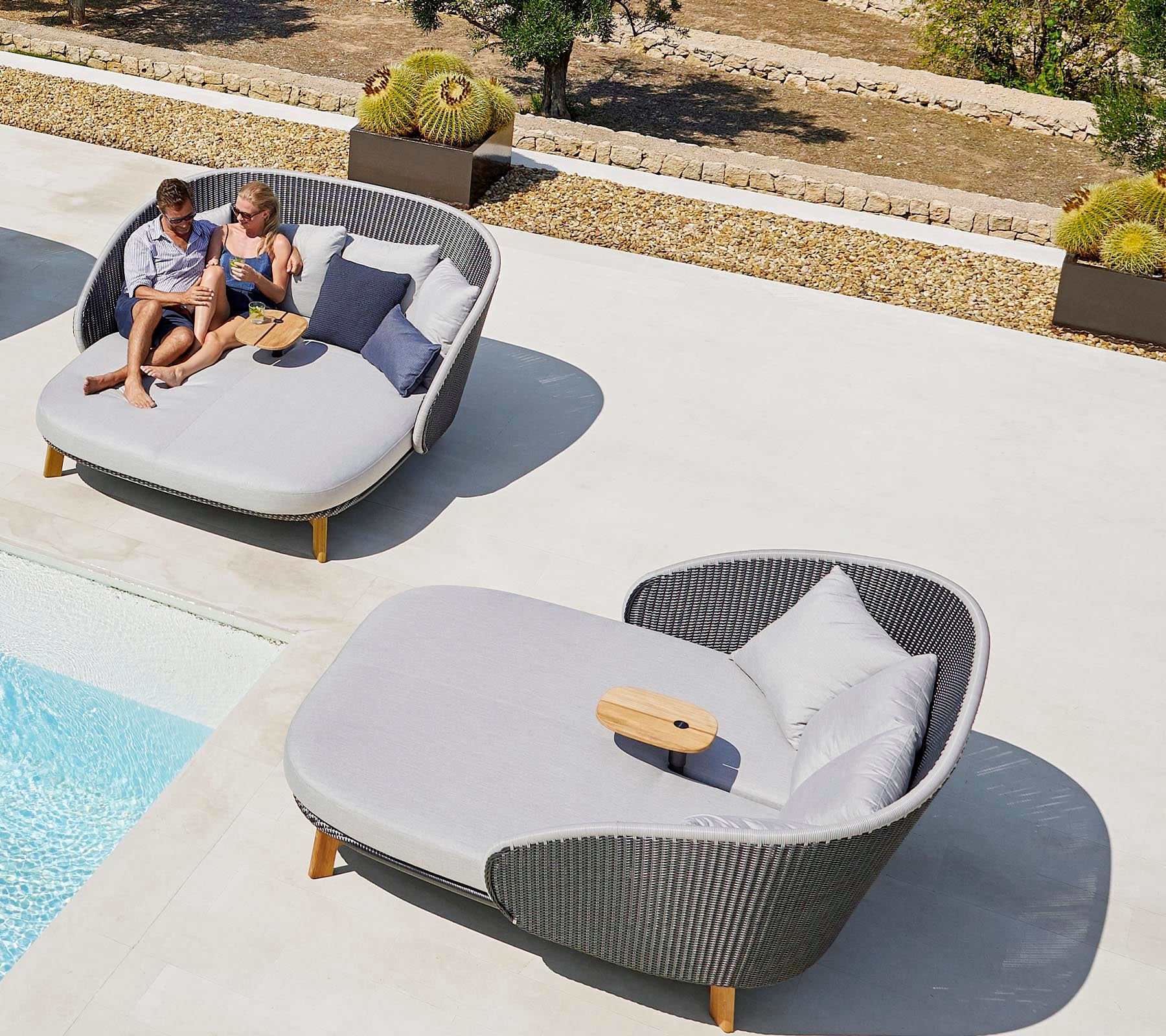 Dos personas se relajan en elegantes tumbonas al aire libre cerca de una piscina, disfrutando de su tiempo libre con cojines y una pequeña mesa.