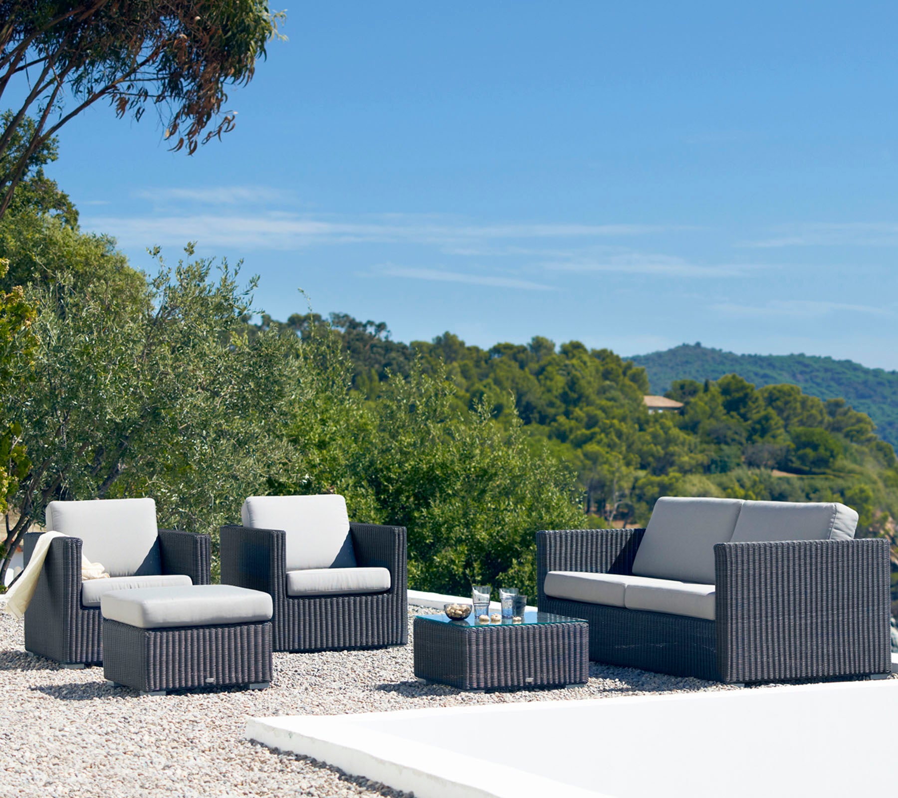 Conjunto de mobiliario exterior elegante que incluye un sofá, dos sillones y una mesa de café con un diseño elegante y cojines neutros, rodeado de vegetación.
