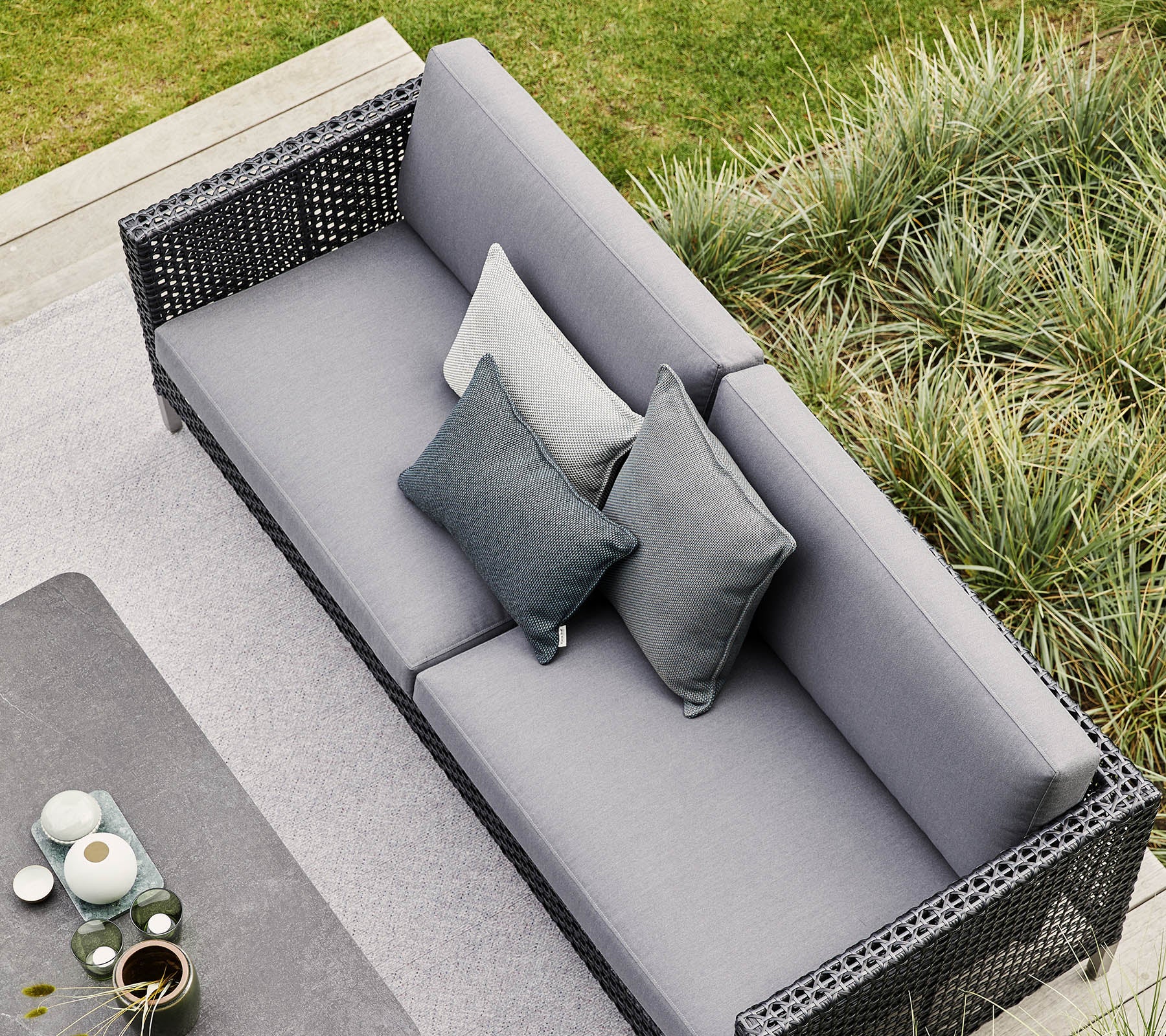 Sofá gris elegante con almohadas decorativas, rodeado de exuberante vegetación y elementos decorativos modernos. Perfecto para relajarse y reuniones.