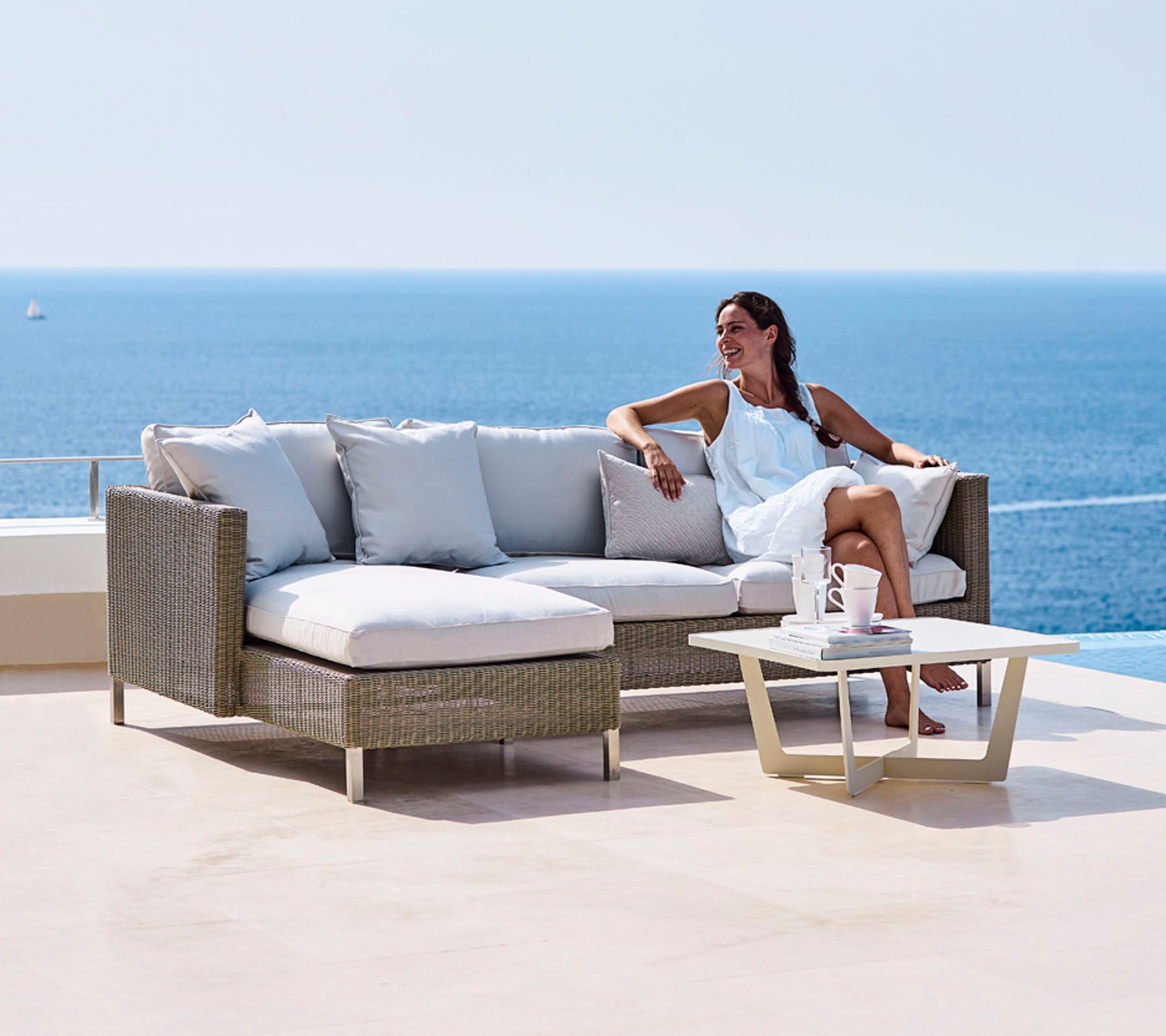 Una mujer se relaja en un sofá elegante con cojines suaves y una pequeña mesa, con vistas a una serena vista del océano.