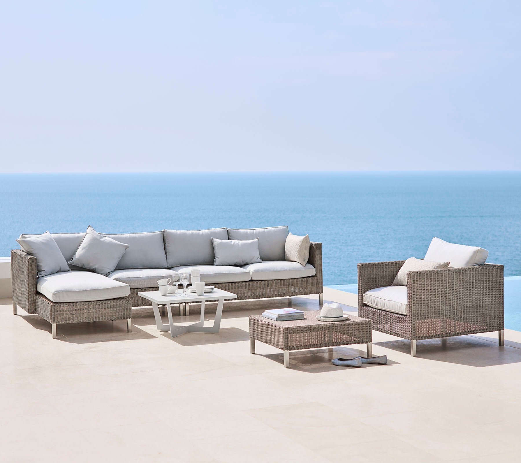 Un elegante arreglo de asientos al aire libre con un gran sofá, una silla y una mesa de café, todos con cojines claros y material trenzado, junto al mar.