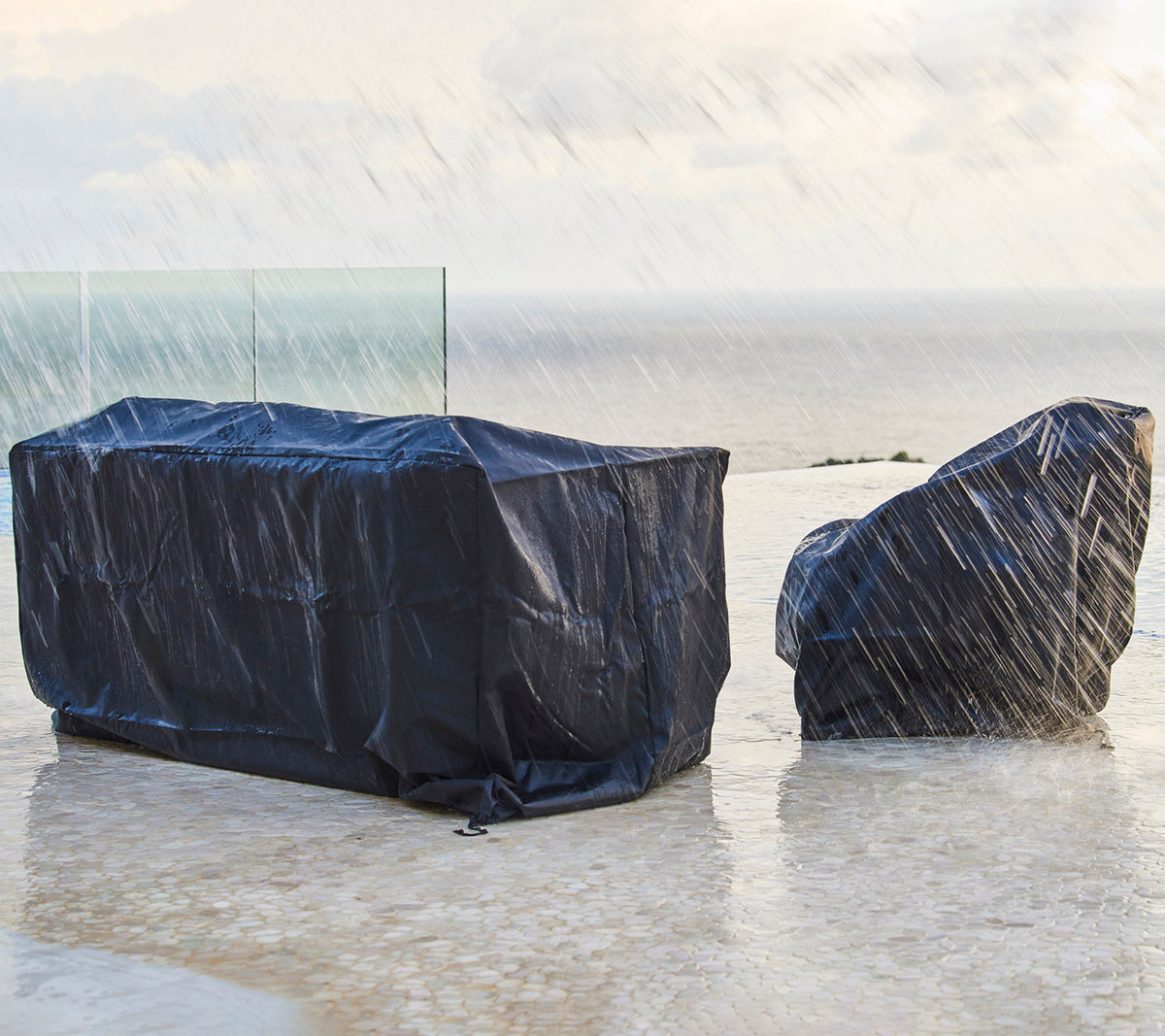 Muebles de exterior cubiertos con fundas protectoras oscuras, a salvo de la lluvia, con un fondo de un cielo nublado y el océano.